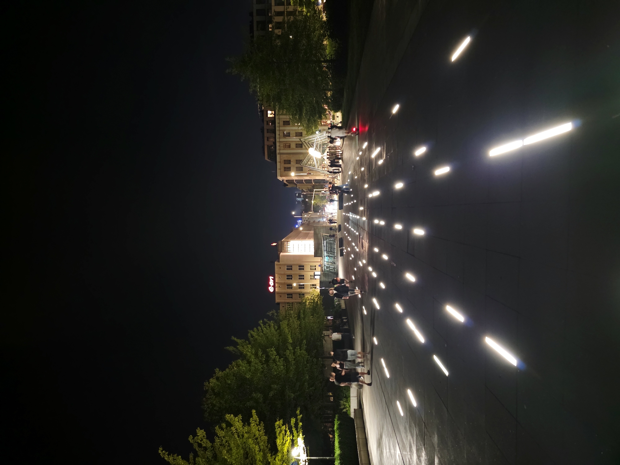 This is a nighttime, vertical shot of a wide pedestrian walkway in Warsaw, Poland. The walkway is paved with dark rectangular tiles and features an array of linear, white lights embedded in the pavement, creating a reflective and illuminated path. Several trees with green foliage are present on the left and right sides of the walkway, some illuminated by external lights.
In the background, there are modern buildings. A prominent building on the left has a red "E.ON" sign. Further down the walkway, people are walking, and a person is riding an electric scooter. The scene suggests a public urban space, possibly a plaza or a wide boulevard, at night.