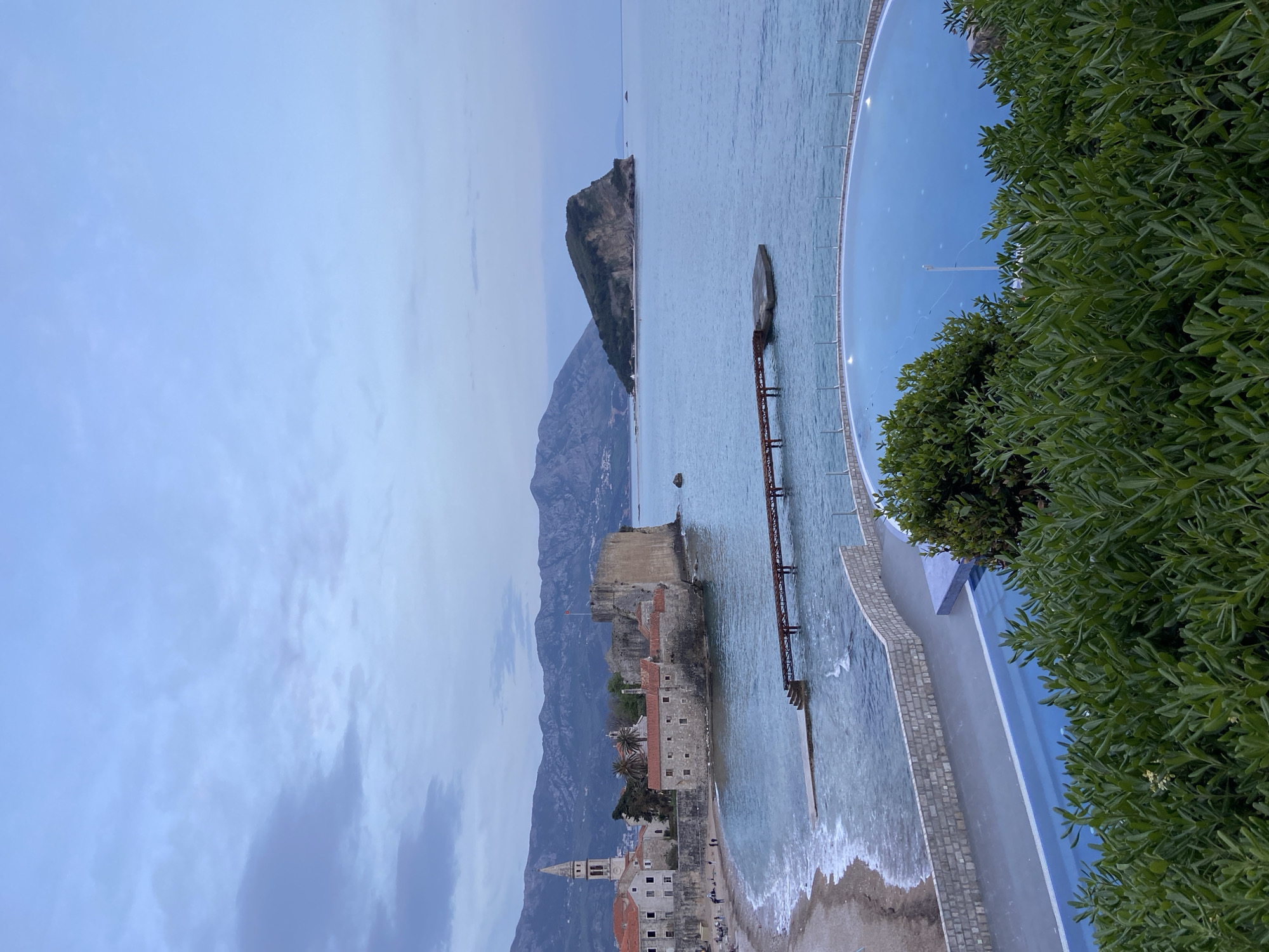 The image shows the old town of Budva, Montenegro. The old town, with its historic stone buildings and a prominent church tower, sits on a peninsula extending into the Adriatic Sea. Behind the town, mountains rise steeply. To the right of the old town, a narrow pier extends into the sea, leading to a small platform. The sea is calm. In the foreground to the right, a curved structure is visible, along with green foliage from trees or bushes. The sky is overcast with clouds.