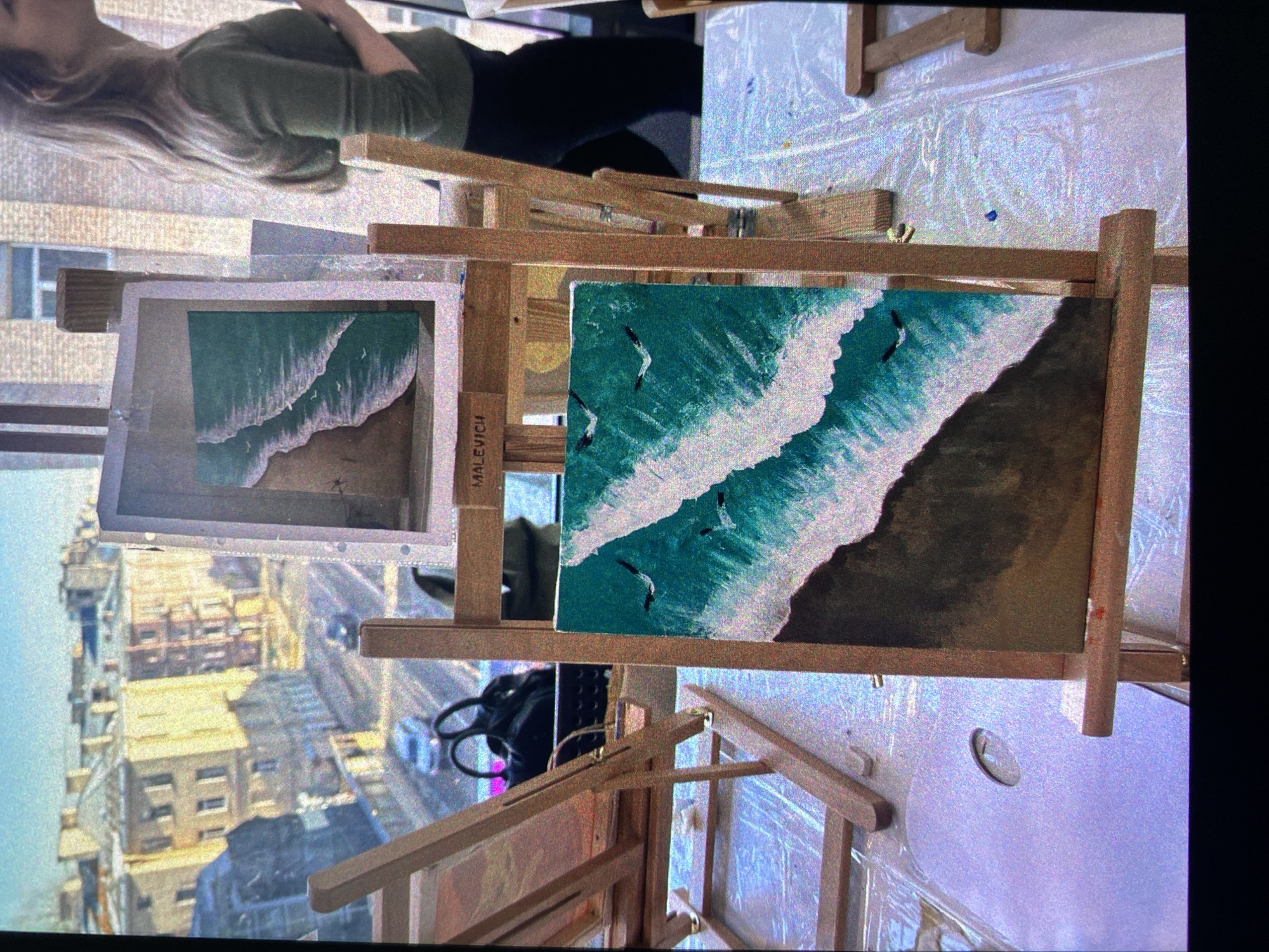 A person is painting a seascape in a room overlooking what appears to be the city of Saint Petersburg, Russia, as evidenced by the architectural style visible through the window. The person, dressed in a green top and dark pants, is slightly out of focus in the background. The focus is on the painting, which features a teal ocean meeting a sandy shore with crashing white waves, and what seem to be seagulls flying above. Another painting, of a similar seascape, is present. Both are propped on wooden easels on a surface covered with protective plastic sheeting. A round, white artist’s palette rests on the surface. A black backpack is on the floor. The room is filled with natural light, which illuminates the scene. The inscription "MALEVICH" is visible on the easel.