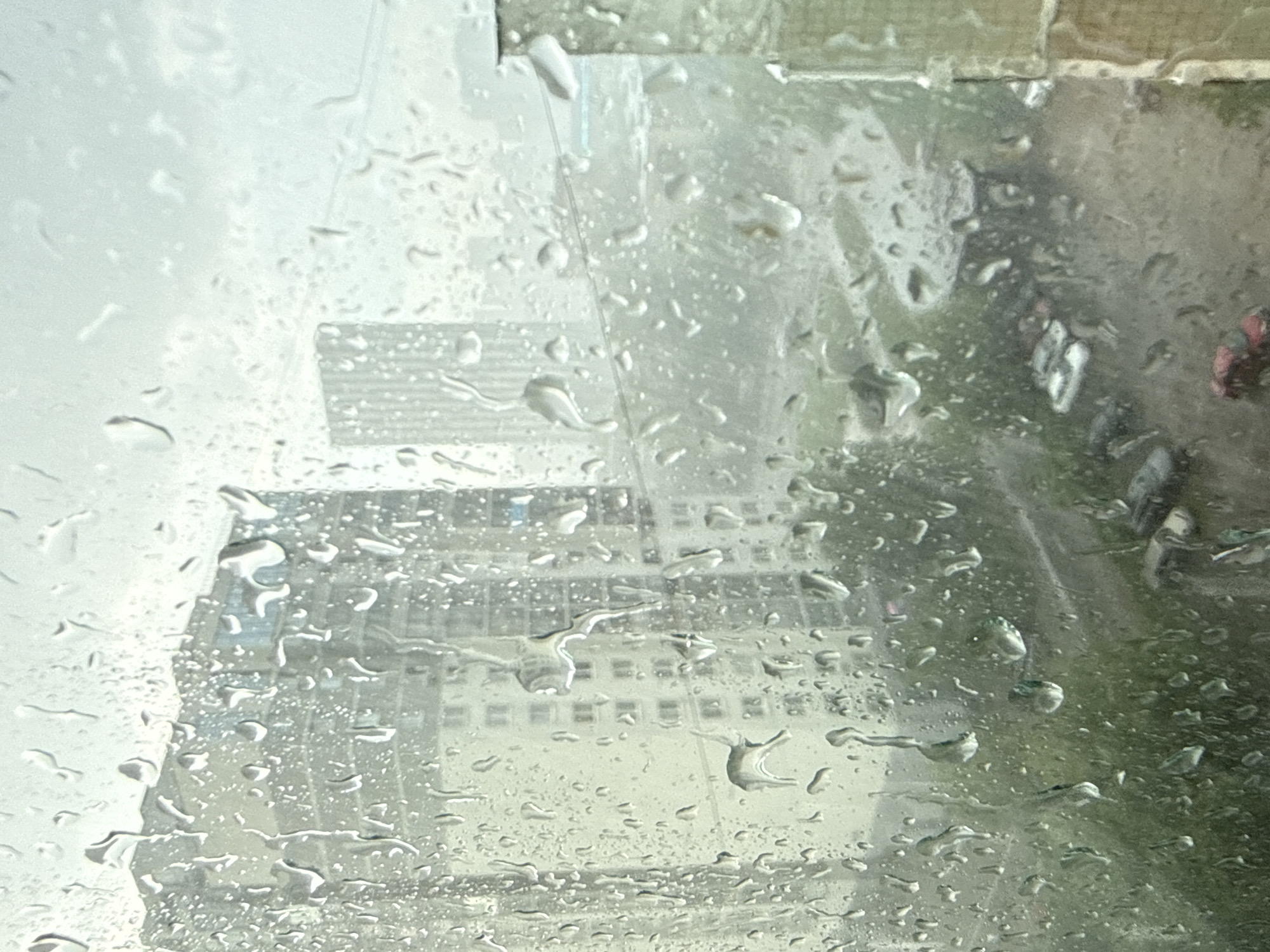 The photograph depicts a rainy scene in Yekaterinburg, Russia. The view is from behind a rain-streaked window, looking out at a cityscape. A tall, modern office building with a grid-like facade is visible in the background. Parked cars line the street on the right side of the frame, partially obscured by the rain. The overall environment suggests an overcast, wet day.