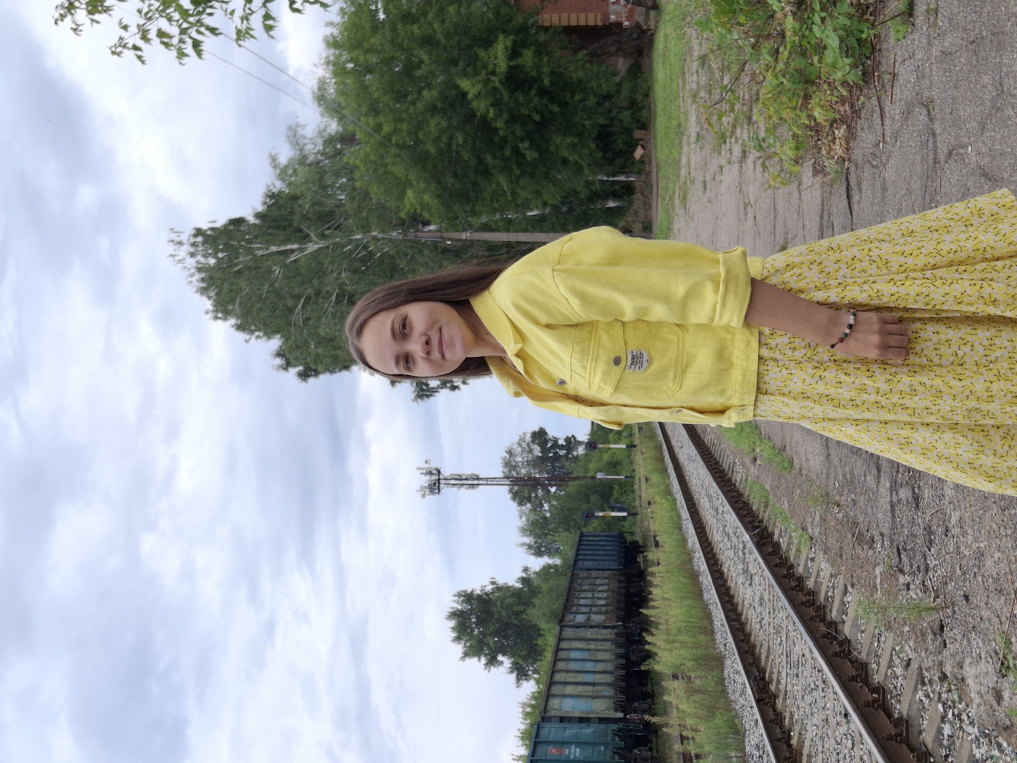 A young woman stands on a platform next to railroad tracks. She is wearing a bright yellow jacket and a yellow floral skirt. In the background, a train of freight cars is stopped on the tracks. Further in the distance, there is a cell tower and trees. The sky is cloudy. This scene is located in Chaikovsky, Russia.