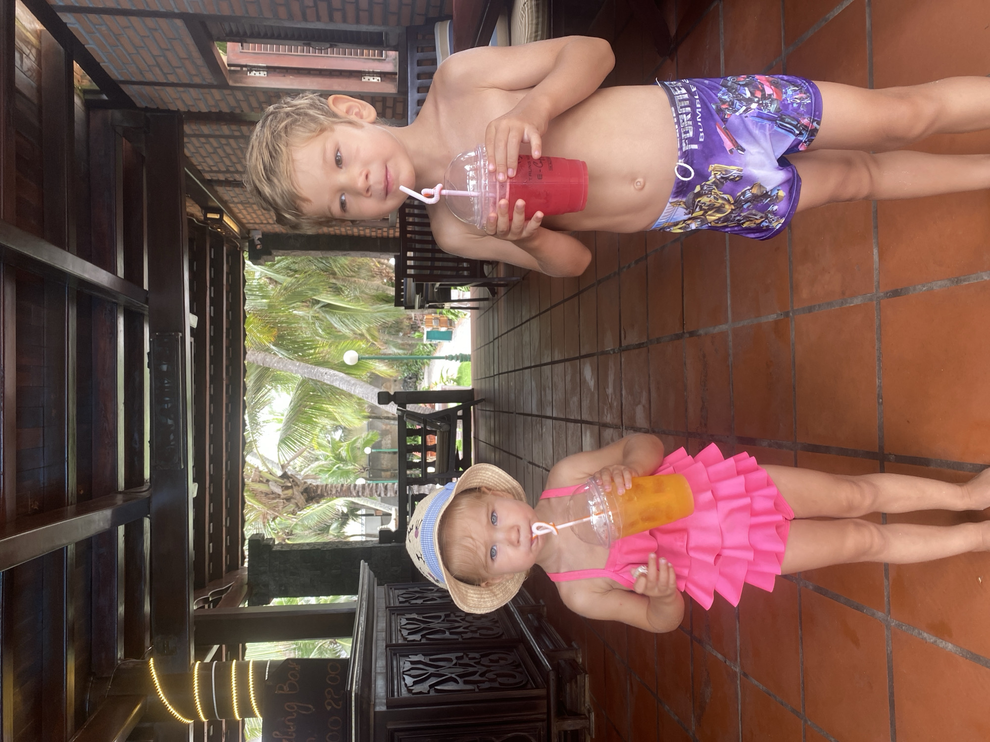Two young children, a girl and a boy, are standing on a tiled floor under a covered walkway in Mũi Né, Vietnam. The girl, on the left, is wearing a pink ruffled swimsuit and a straw hat, and she is drinking from a plastic cup with a straw. The boy, on the right, is shirtless and wearing purple Transformers swim trunks, and he is also drinking from a plastic cup with a straw. In the background, palm trees and a brick building are visible. The overall scene suggests a resort or beachside location.