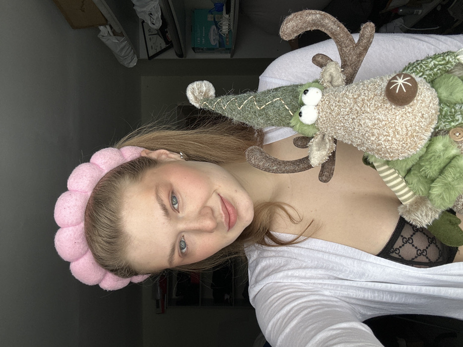 A young woman smiles at the camera, wearing a pink, puffy headband. A stuffed reindeer toy with a green hat and antlers sits atop her right shoulder. She is wearing a white top. A glimpse of a patterned black bra is visible beneath the shirt. The background shows a cluttered shelf and a gray wall. The image is rotated counter-clockwise.