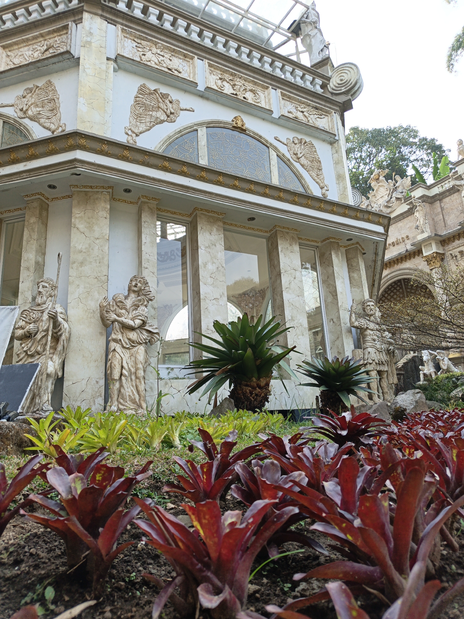 A low-angle, full shot captures a stone building with classical architectural elements and statues in Cibogo, Indonesia. The building features a white facade with beige marble-like accents, golden trim, and intricate bas-relief carvings of winged figures and mythological scenes. Large arched windows with decorative patterns are visible. In front of the building, life-sized stone statues depict figures in draped robes, one holding a staff and the other with outstretched arms. The foreground is filled with a dense planting of red bromeliad plants, with a few spiky green plants behind them and larger tropical plants in the middle ground. The background shows more of the ornate building, trees, and a bright sky. The scene evokes a sense of grandeur and historical artistry.