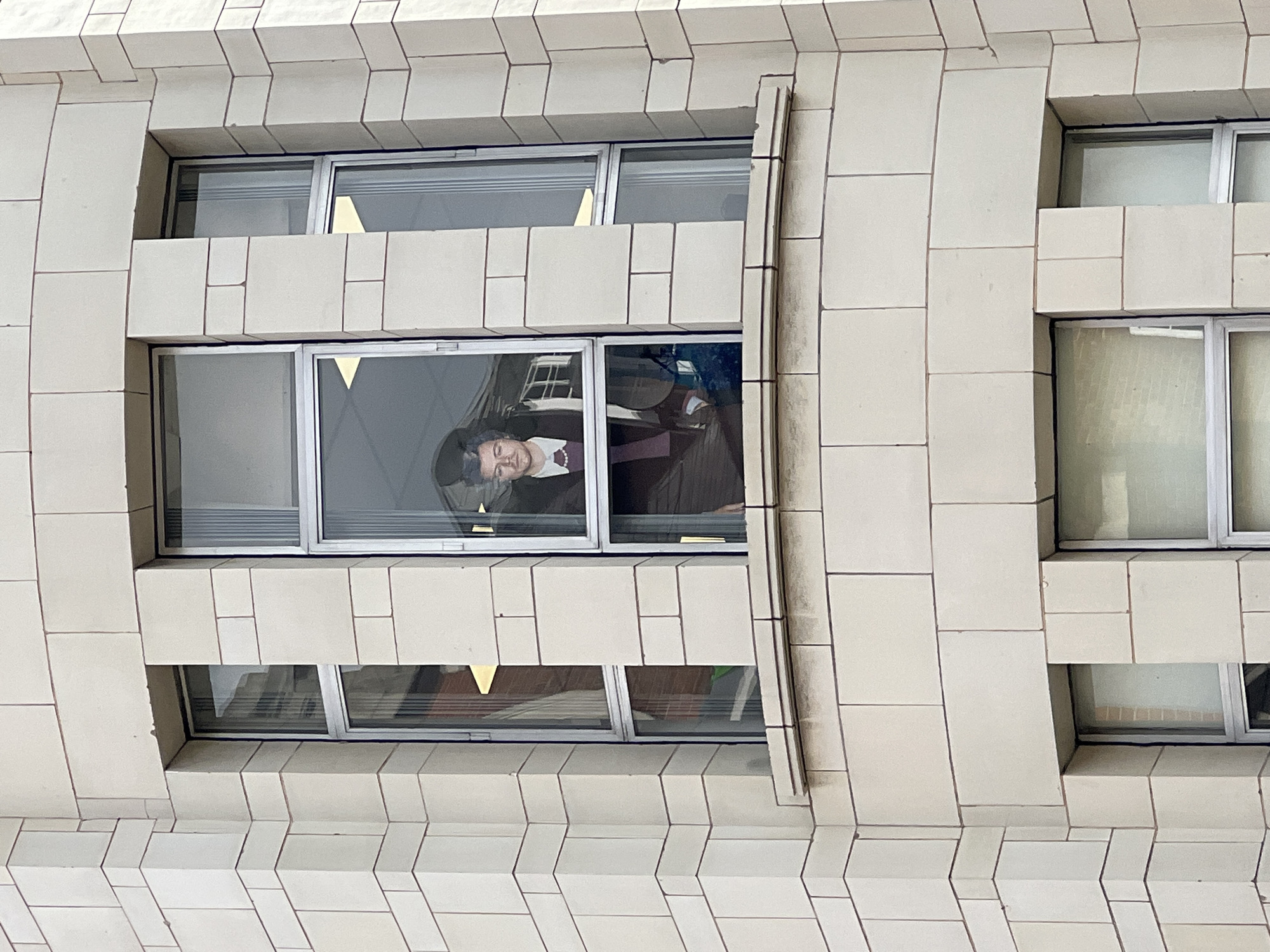 The image presents an upward-angled view of a building's exterior, likely in London, United Kingdom. The facade is constructed from light-colored, rectangular blocks, creating a textured pattern with pronounced horizontal and vertical lines.
Prominently featured is a person's head and upper torso visible through one of the building's windows. The individual has dark hair and appears to be wearing a dark jacket or suit over a white shirt, accented by a decorative element that resembles a pearl necklace or an ornate collar. They are seen looking downwards and slightly to their left, suggesting they are observing something below or standing near the window. Their facial expression is neutral.
The windows feature silver-toned metal frames. Reflections on the glass partially obscure the interior but hint at an office-like setting, possibly showing desks or other furniture. Other windows on the building also exhibit reflections and faint glimpses of interior lighting. The overall scene suggests a modern or commercial urban building. The time of day is not explicitly clear, but the natural illumination points towards daytime. No visible text is present in the image.
