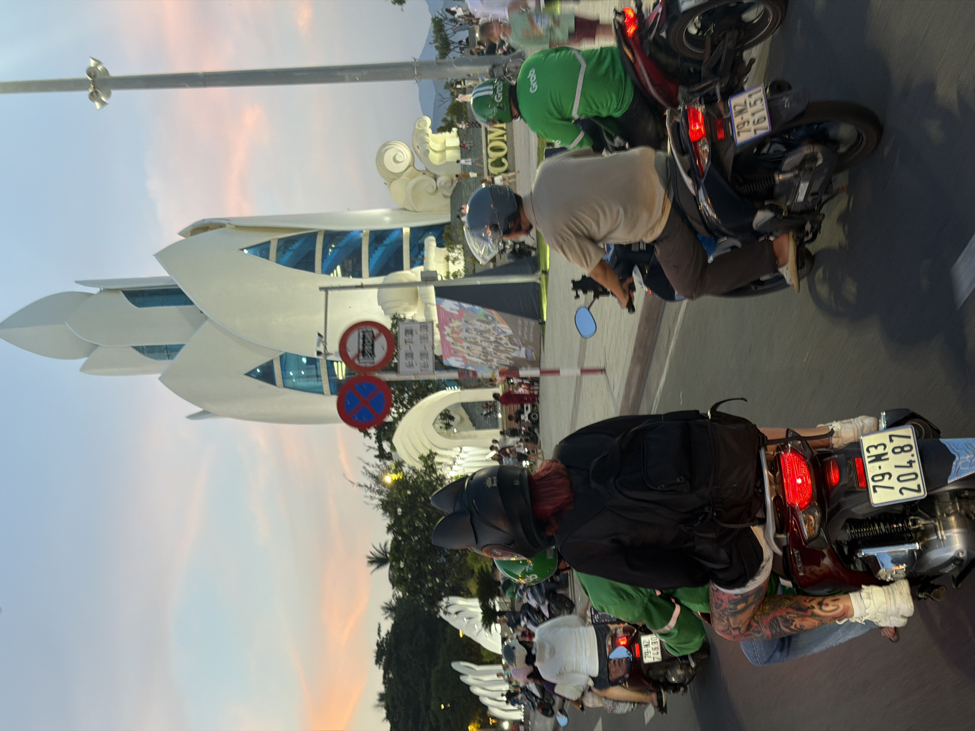 The image captures a street scene in Nha Trang, Vietnam, possibly in Nha Trang Ward, characterized by a distinctive lotus-shaped building in the background. Multiple individuals are riding on motorcycles, navigating the roadway. In the foreground, a person wearing a black helmet with cat ears and a backpack is riding a dark-colored motorcycle. Their legs are heavily tattooed. Another person in a green shirt, presumably a Grab delivery driver, is riding a motorcycle slightly ahead and to the right. A third person in a light-colored shirt is also on a motorcycle to the right of the Grab driver. The sky exhibits a twilight glow with hints of orange and pink clouds. Traffic signs are visible, including a red circular sign with a diagonal bar, indicating a prohibition. The environment appears to be an urban setting with roads, sidewalks, and other vehicles. The time of day suggests late afternoon or early evening.