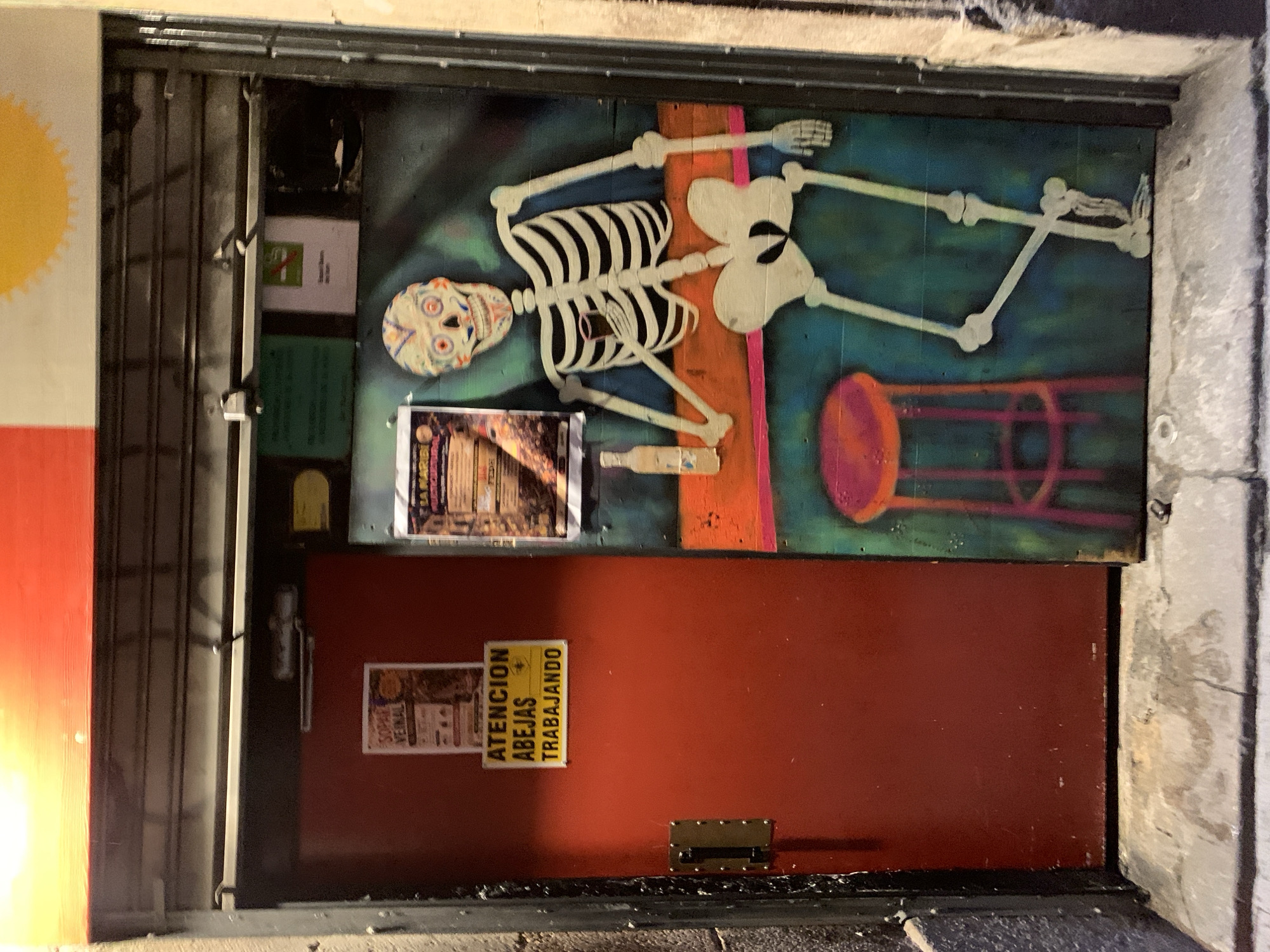 An exterior shot of a closed storefront in what appears to be Barcelona, Spain, is captured at night. The store has a dark red door with a sign in Spanish that reads "Atención Abejas Trabajando" (Attention Bees Working). Above the door, there's a painted mural of a skeleton leaning back on a bar stool, giving the storefront an artistic, perhaps Day of the Dead, vibe. The mural is somewhat worn, and there are a few posters or papers attached to the surface beside it. The storefront is surrounded by what appears to be a metal roll-down security gate, partially open on one side, suggesting the business is currently closed. The street and surroundings are dimly lit, giving the scene a bit of a gritty urban aesthetic.