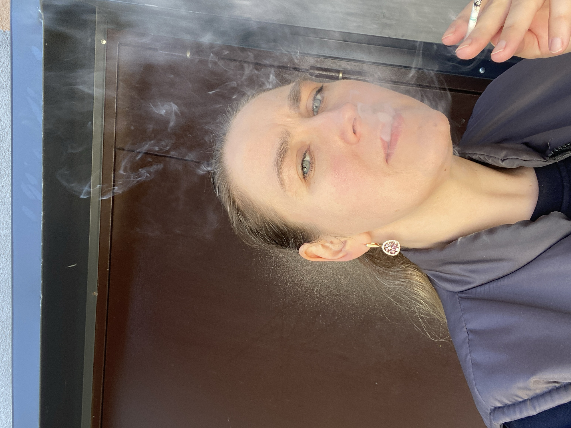 A woman with light hair and eyes holds a lit cigarette, exhaling smoke. She wears a dark jacket and an earring. The background consists of a dark brown surface next to a blue-grey surface. The scene is likely outdoors, although the precise location is not discernible from the image. The city of Yekaterinburg, Russia, cannot be confirmed from this image alone.
