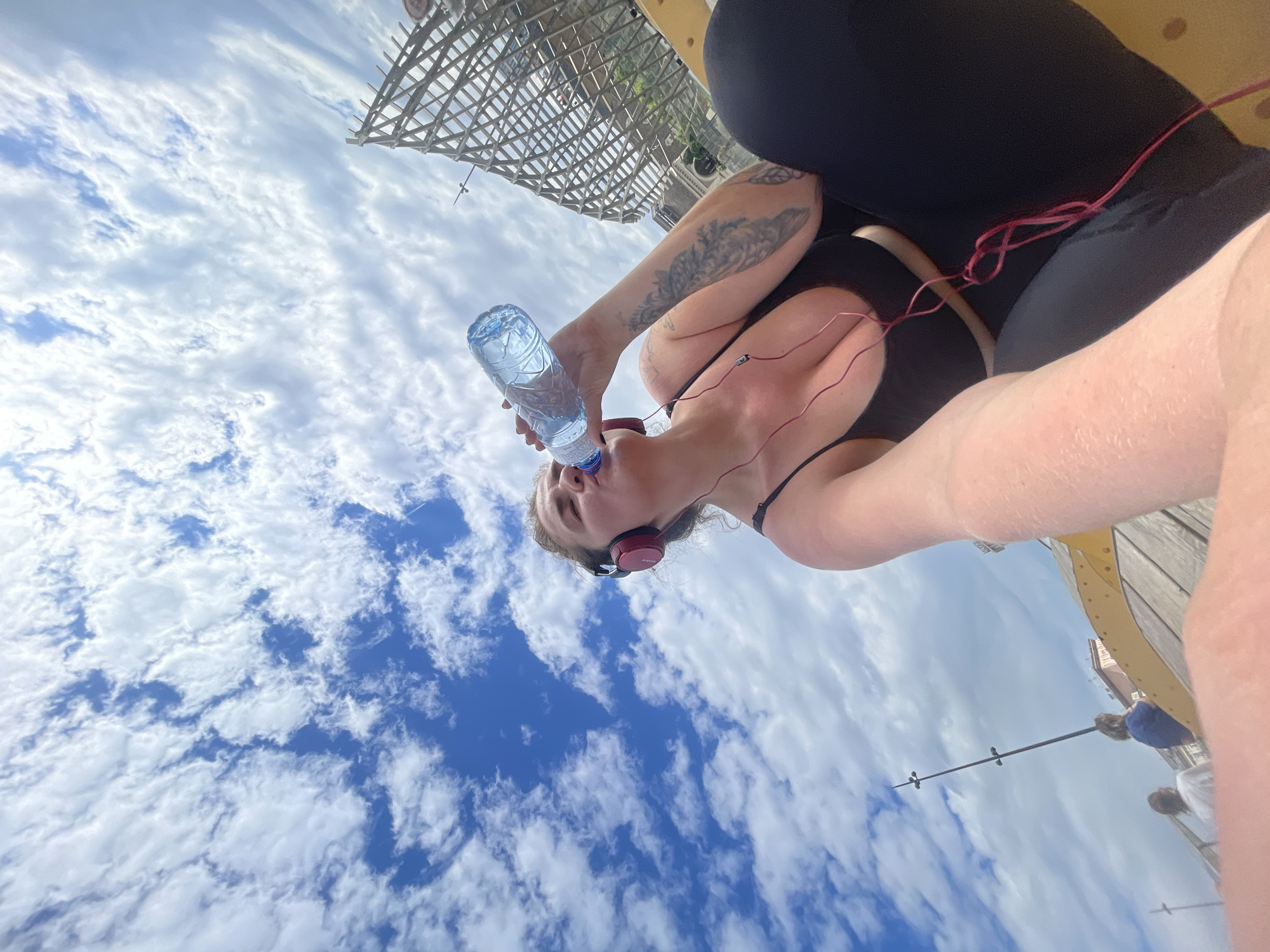 A person is drinking water from a plastic bottle while wearing headphones outside on a partly cloudy day. The person is wearing black leggings and a black top. They have a tattoo on their left arm. The sky is visible behind the person, with many clouds. There is a wooden structure overhead. Other people are visible in the distance. The location is Unknown, Unknown.