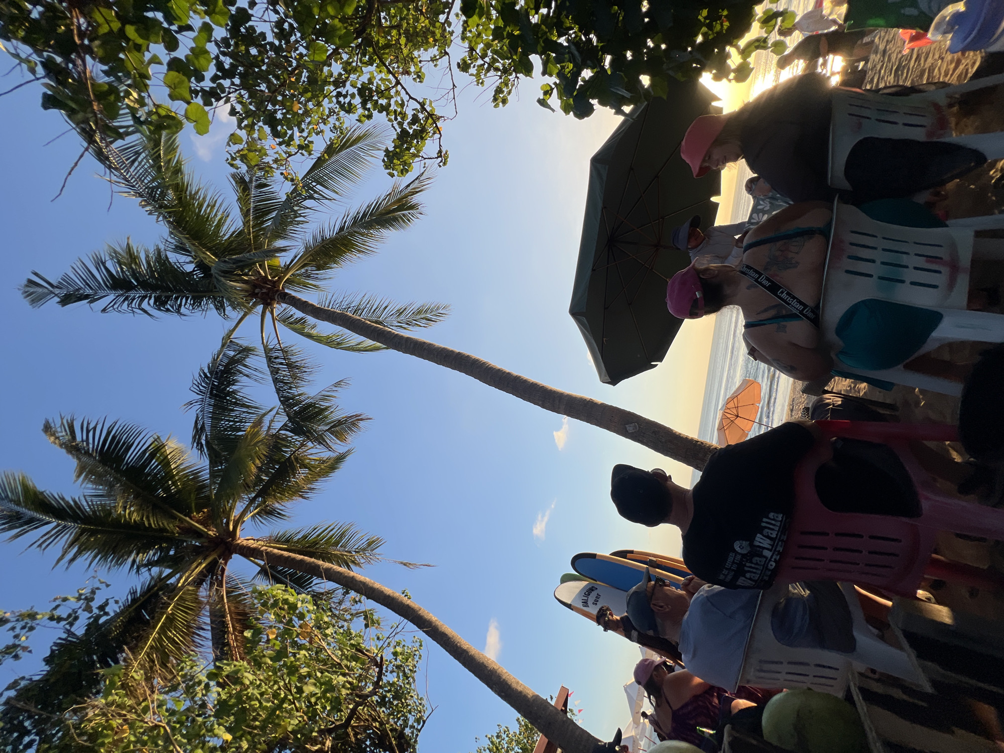 The image captures a scene at Kuta Beach, Indonesia, during the late afternoon. Two tall palm trees frame a clear blue sky with a few wispy clouds. In the foreground, several people are seated on plastic chairs on the sandy beach, facing towards the ocean. The individuals appear to be a mix of locals and tourists, dressed in casual beach attire. Some are looking out at the sea, while others are engaged in conversation.
Behind the seated individuals, surfboards stand upright, hinting at the beach's popularity for watersports. A large, dark green beach umbrella provides shade for some of the group. In the distance, the ocean is visible, with gentle waves rolling in. The setting sun casts a warm glow on the water and the beach, creating a serene atmosphere.
To the left of the frame, a vendor appears to be setting up or selling goods, with several white beach umbrellas visible in the background. The overall impression is one of a relaxed evening gathering at a popular beach destination.