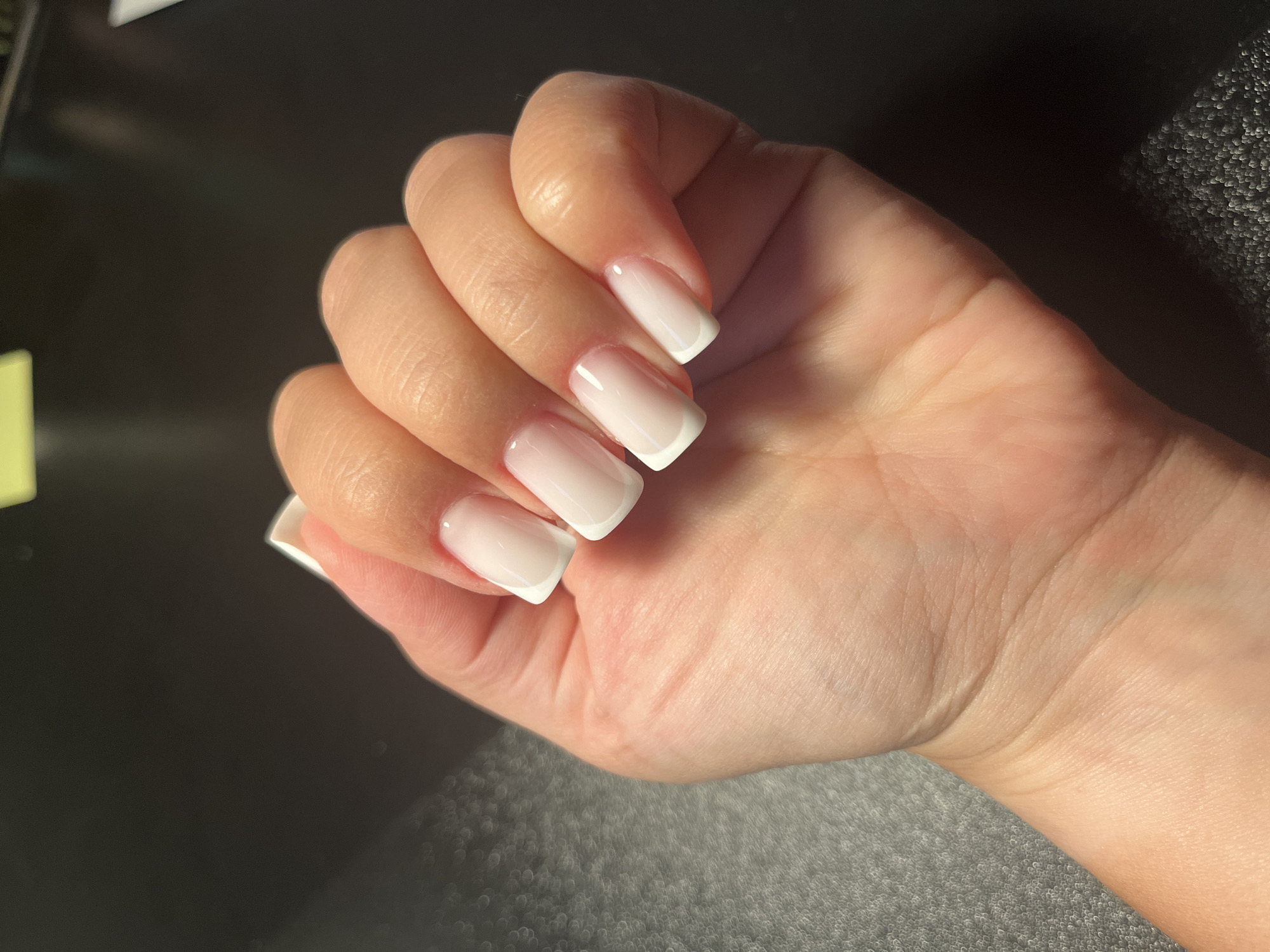 A close-up, high-angle shot shows a person's left hand, fingers extended, against a dark, textured surface. The nails are manicured with a classic French tip style, featuring a light pink base and a crisp white tip. The skin on the hand appears smooth with visible texture from pores and fine lines. The background is dark and out of focus, with some areas showing a subtle bokeh effect, possibly from a patterned surface or lighting. The lighting is bright and comes from the top left, creating gentle highlights on the nails and skin. The image does not contain information about the city of Maardu, Estonia.