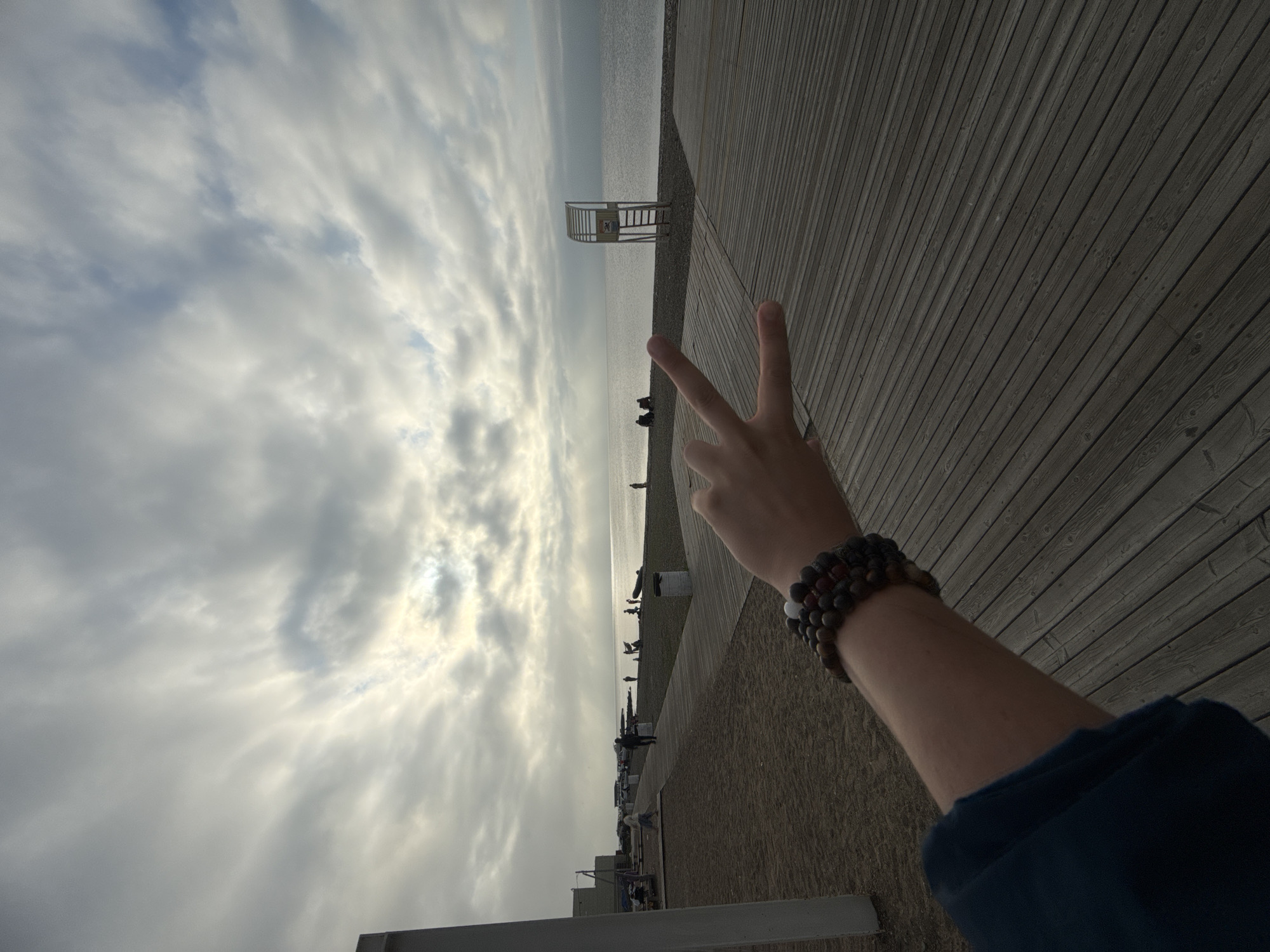 A person's left arm, adorned with several bracelets, is extended towards the camera, with the hand forming a peace sign. The arm is covered by a dark blue sleeve. The background depicts a beach and the sea under a cloudy sky. In the distance, people are visible walking and sitting on the beach. A lifeguard tower stands near the water's edge. The foreground is dominated by a wooden boardwalk. The scene appears to be set in Vladivostok, Russia, likely during a time of moderate weather.