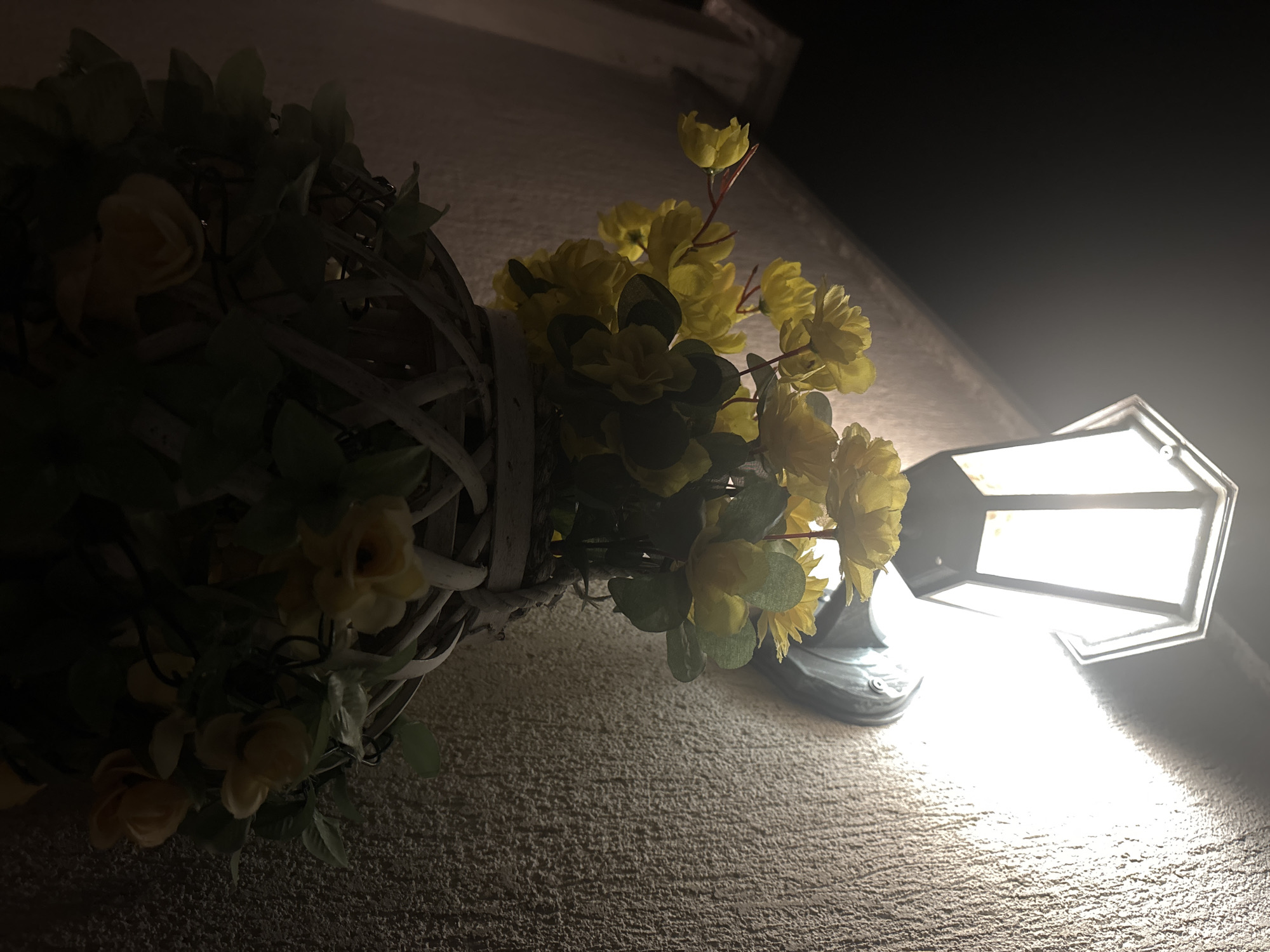 A dim, low-angle indoor shot focuses on a lit, rectangular lantern attached to a textured, cream-colored wall. To the left of the lantern, a wicker basket overflowing with yellow flowers and green leaves is mounted on the wall. The lantern illuminates the flowers and the wall immediately beneath it. The background is dark, suggesting a nighttime setting. No people are visible. There are no elements suggesting the city of Lom, Bulgaria.