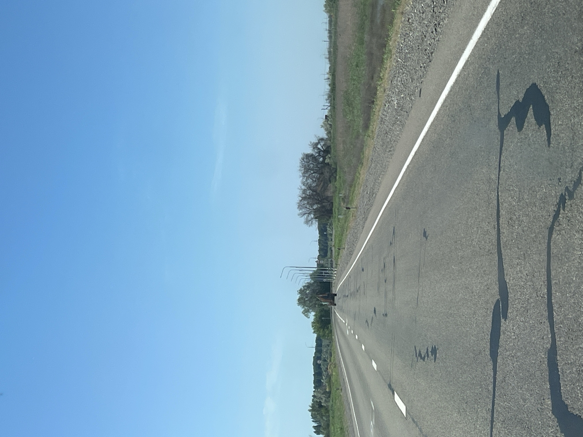 This is a forward-facing, eye-level, vertical, medium shot of a two-lane asphalt road in Bakhtemir, Russia. The road is clear of traffic except for a single excavator in the distance. The sky is clear and blue. On the right side of the road, there is a ditch with tall grass and reeds. On the left side of the road, there are trees. The road has white dashed lines on the left and a solid white line on the right. There are dark patches on the asphalt, possibly water or oil stains, and some shadows cast on the road. The excavator is facing away from the camera and appears to be stationary. There are also streetlights visible along the right side of the road.