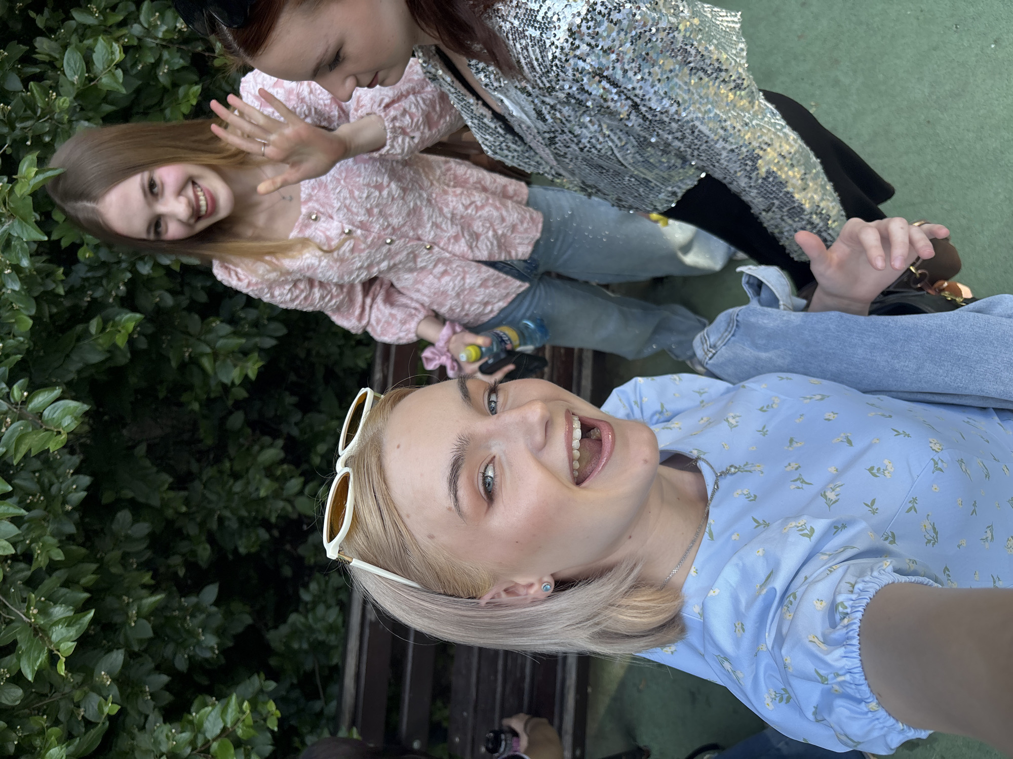 An outdoor, eye-level group selfie captures three young adult women in a public green space in Vsevolozhsk, Russia, under bright daylight.
In the immediate foreground, a blonde woman with white-framed sunglasses pushed onto her head smiles broadly, mouth open, looking directly at the camera. She wears a light blue short-sleeved top with a white and yellow floral pattern, light blue jeans, a silver chain necklace, and a small blue stud earring in her left ear. Her right arm is extended, holding the camera, with a partially visible left arm bent at the elbow, gripping a brown object.
Behind her and slightly to the right, a second woman with long, light brown hair smiles, her left hand raised in a waving gesture. She wears a textured pink long-sleeved blouse with a square neckline and a pattern of raised fabric, paired with light blue jeans embellished with small silver reflective spots. Her right hand holds a clear plastic water bottle with a yellow cap, and a pink scrunchie is on her wrist.
To the right of the second woman, a third woman with reddish-brown hair, secured with a black hair accessory, is visible from the chest up, facing slightly left. She wears a silver sequined jacket over a black top. Her expression is a faint smile.
The background consists of dense, dark green foliage from bushes or trees, with some small white blossoms or berries. A dark brown slatted wooden bench is partially visible behind the women. The ground in the lower right is a light green, flat surface. A blurry figure is partially visible to the far left, behind the foreground woman, holding a black cylindrical object.