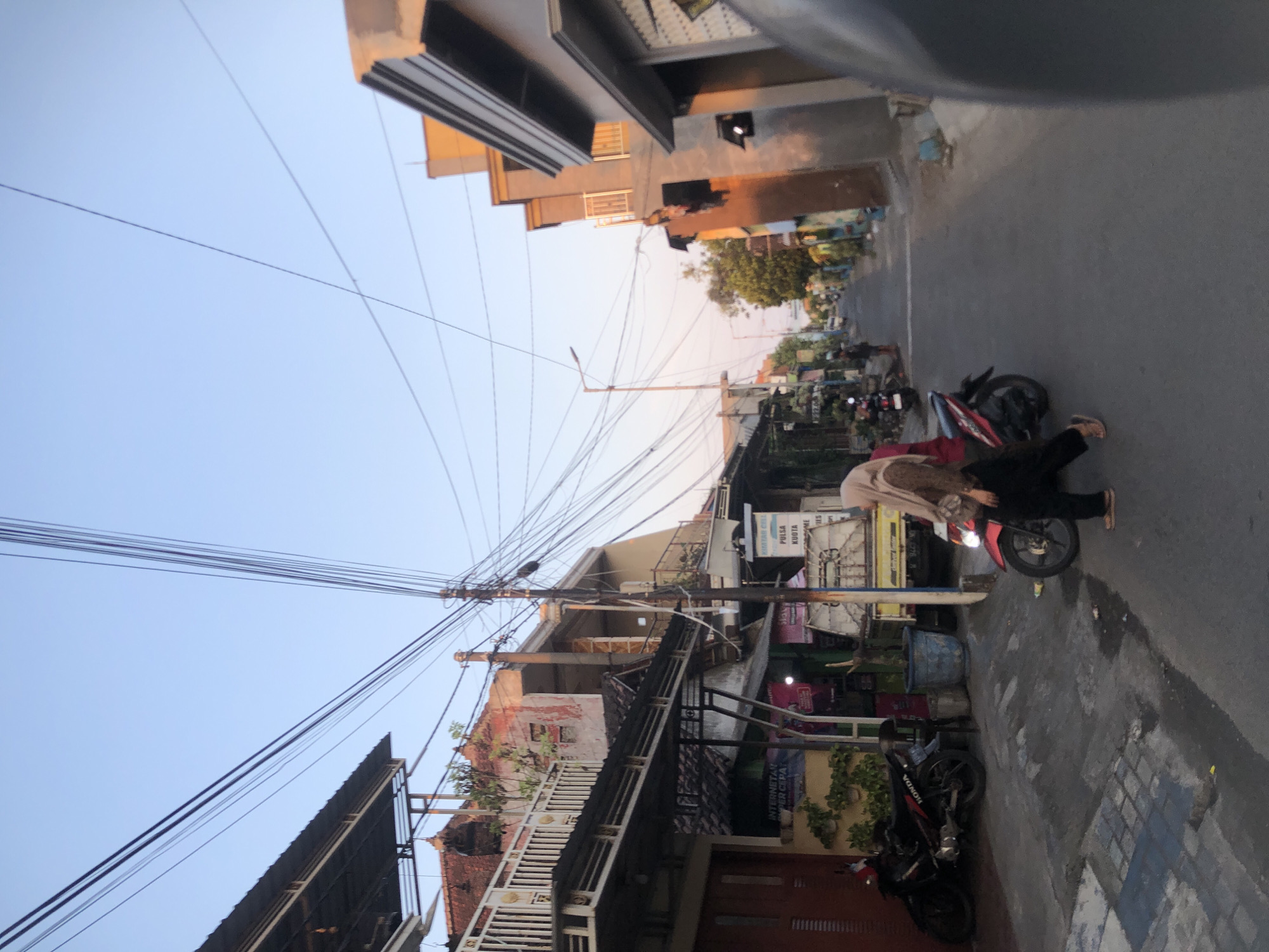 In Tenggulunan, Indonesia, a street scene unfolds under a clear sky. The perspective is from a low angle, possibly from a vehicle, looking down a narrow street lined with buildings and overhead electrical wires.
On the left, a two-story building with a white railing on the upper floor is visible, its lower section featuring a red door. Next to it, a black Honda motorcycle is parked. Further down the street, a smaller stall with signs in Indonesian can be seen.
In the center of the street, a woman wearing a hijab and a brown patterned top over black pants walks beside a red and black motorcycle. She is moving away from the viewer. Another person is standing further down the street, a silhouette against the receding road.
The buildings on the right are modern, with one having multiple stories and balconies with white railings, illuminated by the setting sun which casts a warm glow. Another building features a gray facade with dark window frames.
The street surface is asphalt, with some patchy gray areas and a slight sheen suggesting moisture. The overall impression is of a residential or commercial street in a populated area, with the dynamic overhead network of wires being a prominent feature.