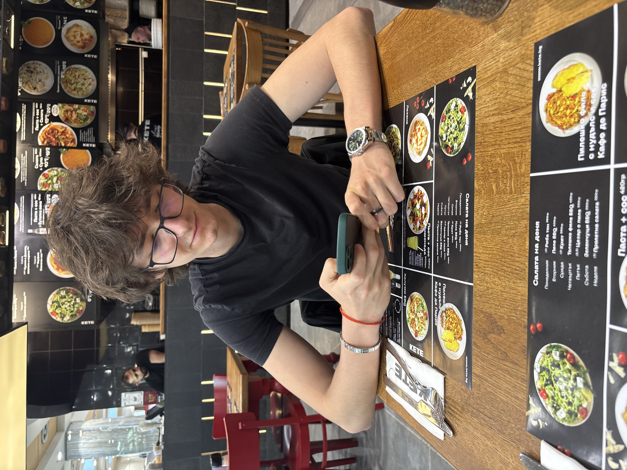 A young man with brown hair and glasses is seated at a wooden table in a restaurant. He is wearing a black t-shirt and has a watch on his left wrist and bracelets on his right wrist. He is holding a green smartphone. On the table is a menu with food items listed in Bulgarian and English, as well as cutlery and condiments. The menu features the restaurant's name, "KETE," and the restaurant's website. Red chairs are visible in the background. Based on the menu and language, the location is Sofia, Bulgaria.
