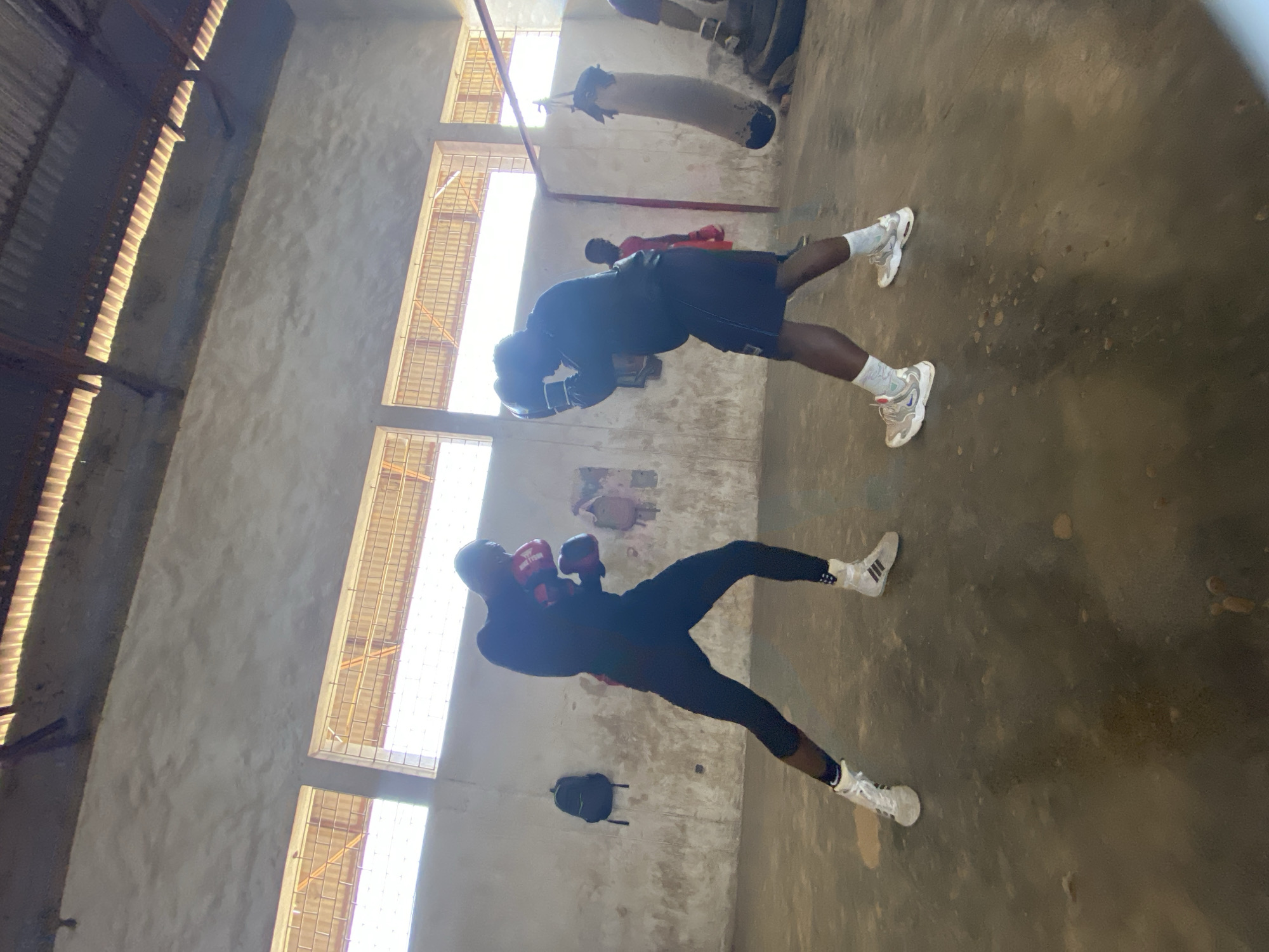 The image shows two boxers training indoors in a gym in Jos, Nigeria. The gym appears to be a basic, older facility with barred windows allowing natural light in. The walls are a faded, light color. The boxers are dressed in athletic wear and boxing gloves. One boxer has red gloves and the other has black. They are engaged in shadow boxing, practicing their punches and footwork. Other gym equipment, including a punching bag, is visible in the background. The floor appears to be concrete.
