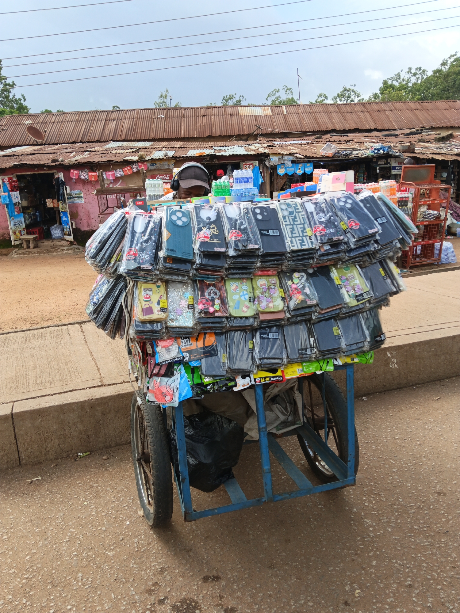 A street vendor displays an abundance of colorful phone cases on a cart. The cart, constructed of blue metal, has two large spoked wheels. The phone cases, neatly arranged on multiple racks, feature various designs and colors, including black, white, green, and patterned cases. Some cases have clear packaging, revealing intricate designs, while others are solid colors. At the bottom of the cart, accessories like a USB car charger and orange earphones are visible. The vendor, partially obscured by the cart, is wearing headphones and a cap, suggesting a busy marketplace environment.
Behind the cart, a modest shop with a corrugated metal roof is visible. The shop appears to sell a variety of items, including bottled drinks like Coca-Cola and water, as indicated by the visible labels. There are shelves stocked with products, and some items are displayed outside the shop. The ground is a dirt and gravel mixture, typical of an outdoor market. The sky is overcast, suggesting a cloudy or possibly rainy day. The overall scene depicts a vibrant, everyday market setting where a vendor is actively selling their wares. Visible text includes "Coca-Cola," "POS," "BYTE PRO," "KISS," "DESIGN," "FAST CHARGING," "USB CAR CHARGER," and "AL-DI ALBARICA."