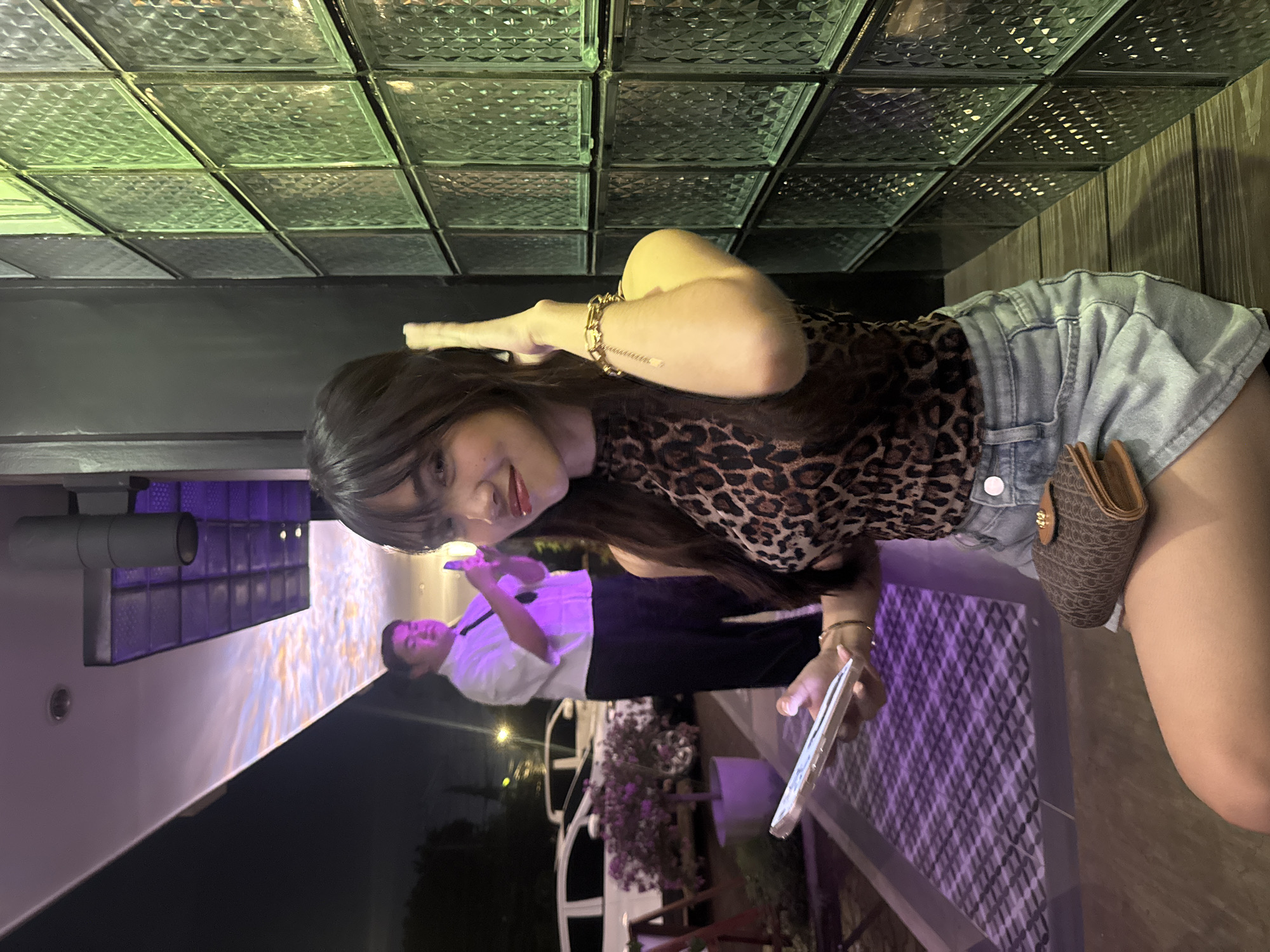 A young woman with dark hair and bangs is posing for a photo at night in what appears to be an outdoor seating area of a restaurant or bar. She is wearing a leopard print halter top and denim shorts, with a small brown clutch purse on her lap. She is holding a smartphone in her left hand and touching her hair with her right hand. In the background, a man in a white shirt and black tie is also holding a smartphone, appearing to take a picture. The environment features a patterned tiled floor, a dark wall, and a wall of textured glass blocks illuminated with green light. In the further background, there are cars and trees, suggesting an urban setting. The ceiling above the man displays a projection of a colorful sky. The overall lighting suggests it is nighttime, with artificial lights creating a vibrant atmosphere. This scene is situated in Davao City, Philippines.