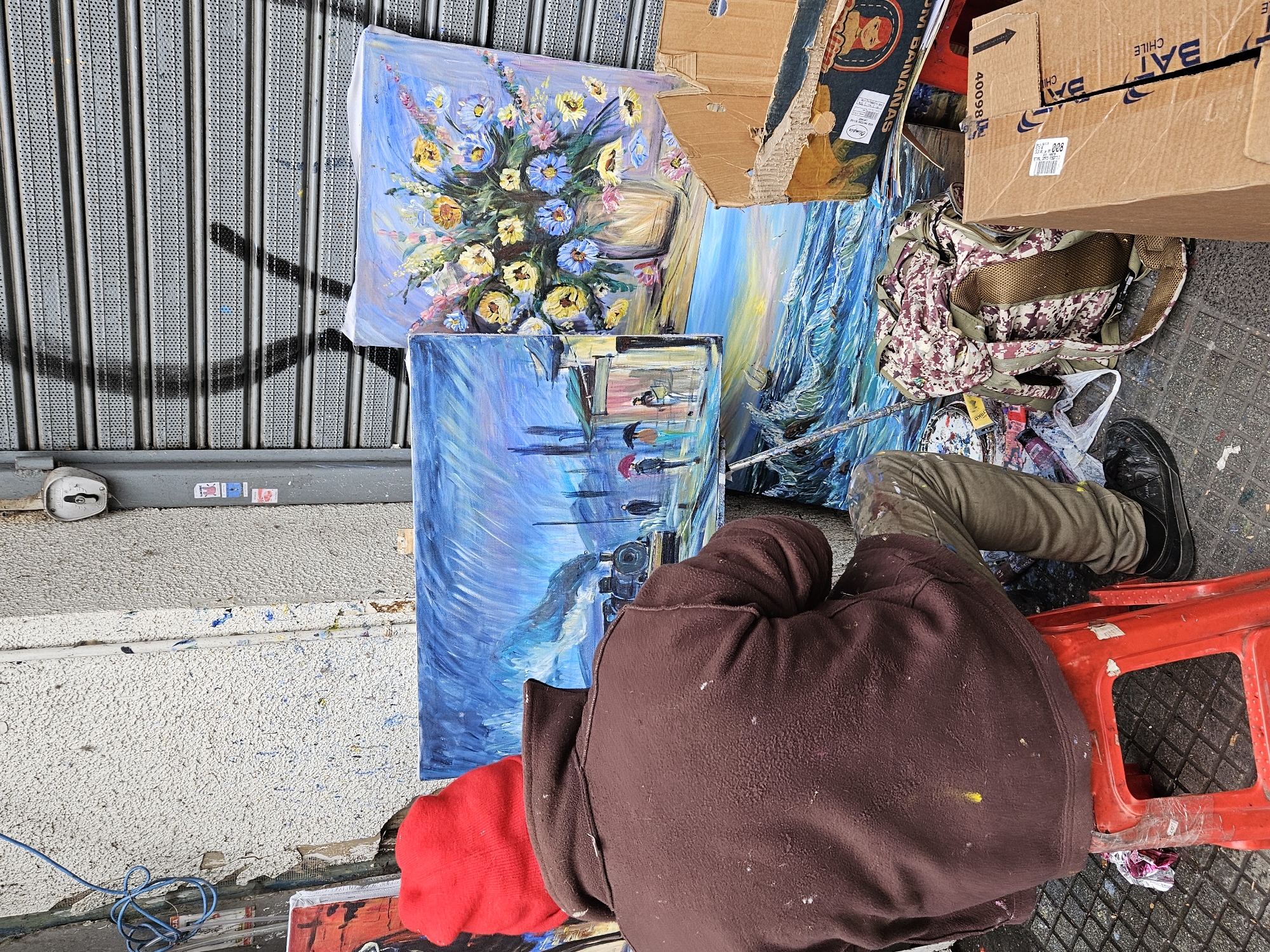 The image shows an artist sitting on a red plastic stool on a sidewalk, painting on the street in Concepcion, Chile. He is wearing khaki pants with paint stains, a brown hooded sweatshirt, and a red beanie. His black shoe is visible. Four paintings are arranged next to him. One depicts a vase of colorful flowers, another a seascape, and the remaining two display depictions of buildings and objects. Paint tubes, brushes, and a palette are scattered nearby. A cardboard box, a backpack, and other discarded items are also present on the sidewalk. The background shows a corrugated metal wall with shadows cast on it.