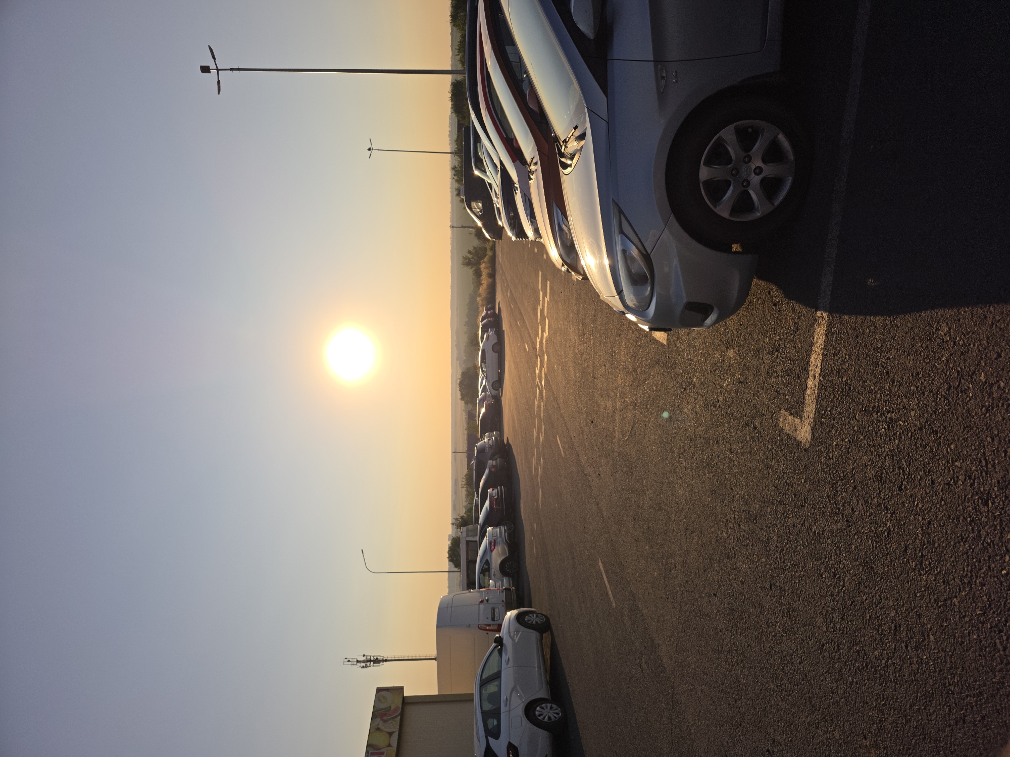 A view of a parking lot at sunset in Rostov-on-Don, Russia. The sun is low on the horizon, casting a warm glow across the scene. Several cars are parked in the lot, and a building with some signage is visible on the left. A cell tower and a street lamp are also present. The ground is asphalt, and parking lines are visible. The sky is clear with a gradient of blue and orange.