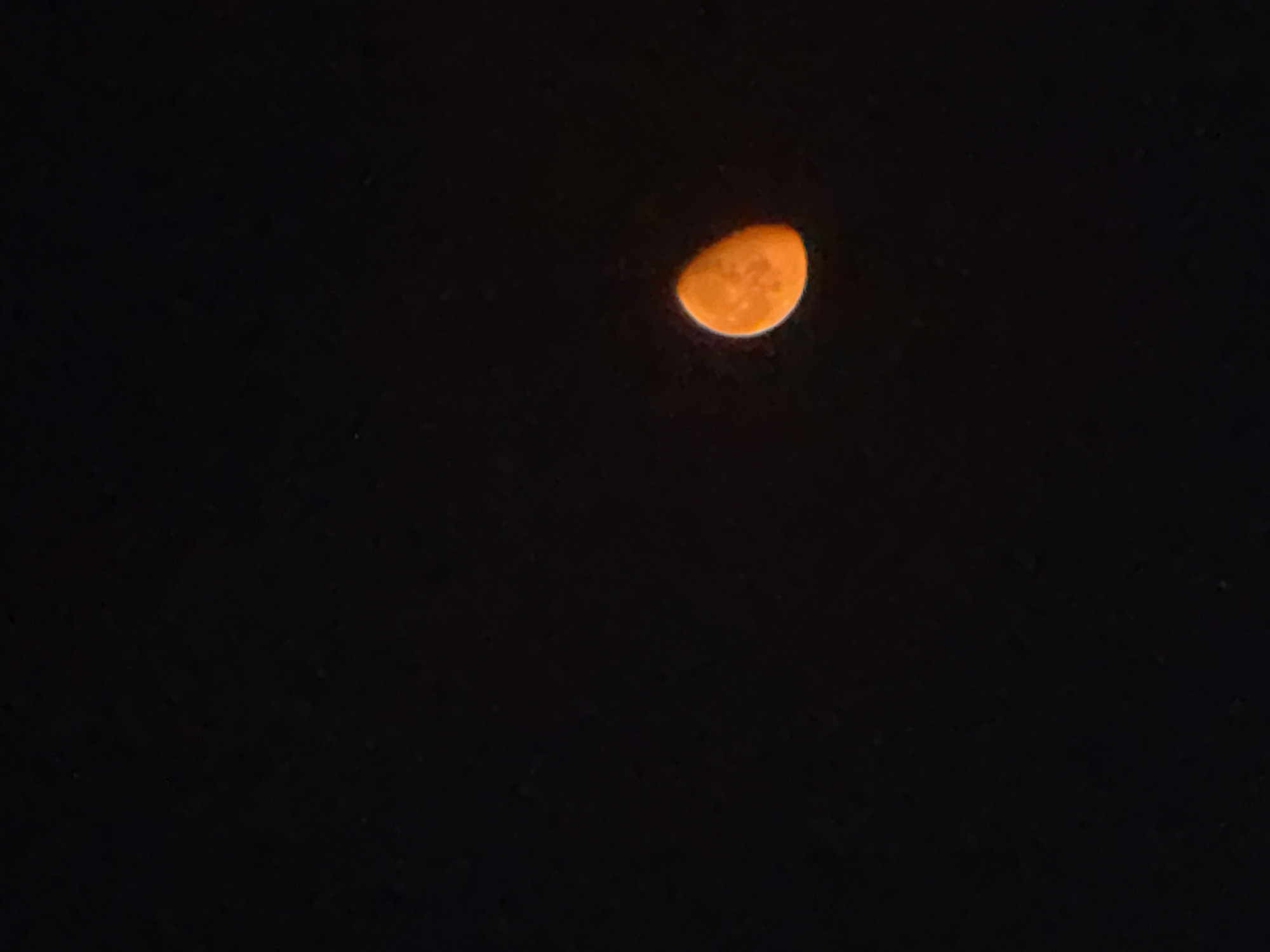 The image shows a crescent moon in the night sky. The moon is orange in color and appears to be illuminated from the right. The background is a dark, black sky. There are no other visible celestial bodies or objects in the image. The location of the city of Tbilisi, Georgia, is not identifiable from the image.
