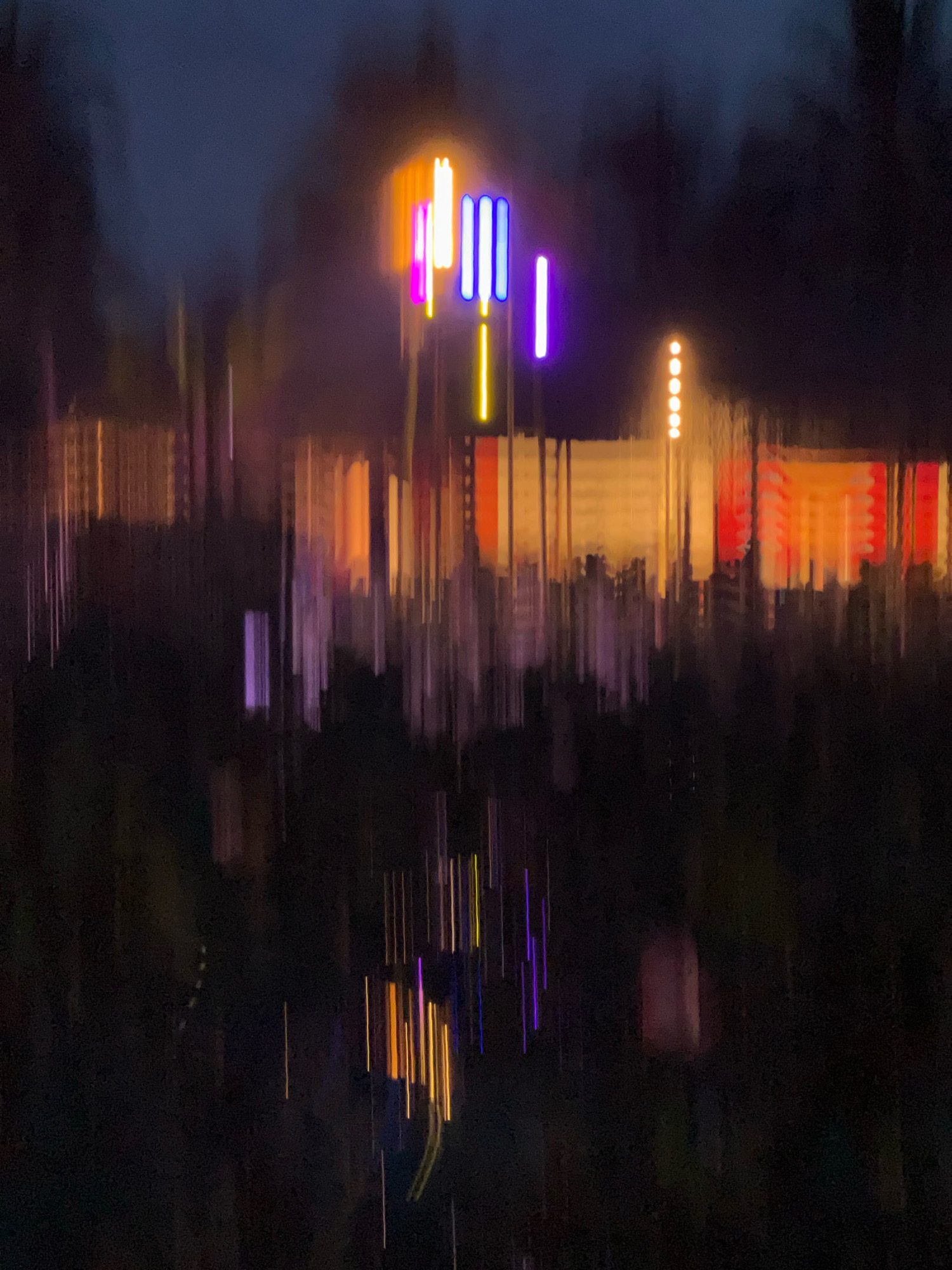 The image is an abstract, blurred depiction of lights at night, likely in Nha Trang, Vietnam, given the provided context. The scene is dominated by vertical streaks of color, suggesting movement or long exposure photography. Prominent are bright, neon-like vertical lights in shades of pink, purple, yellow, and blue, arranged in clusters. Behind these, elongated bands of orange, yellow, and red lights are visible, resembling illuminated windows or signage on buildings. The overall impression is one of a vibrant, possibly urban, nighttime environment, with the blurriness obscuring specific details of any people or structures, creating an impressionistic effect. The lower portion of the image is dark, with faint vertical reflections or light trails.