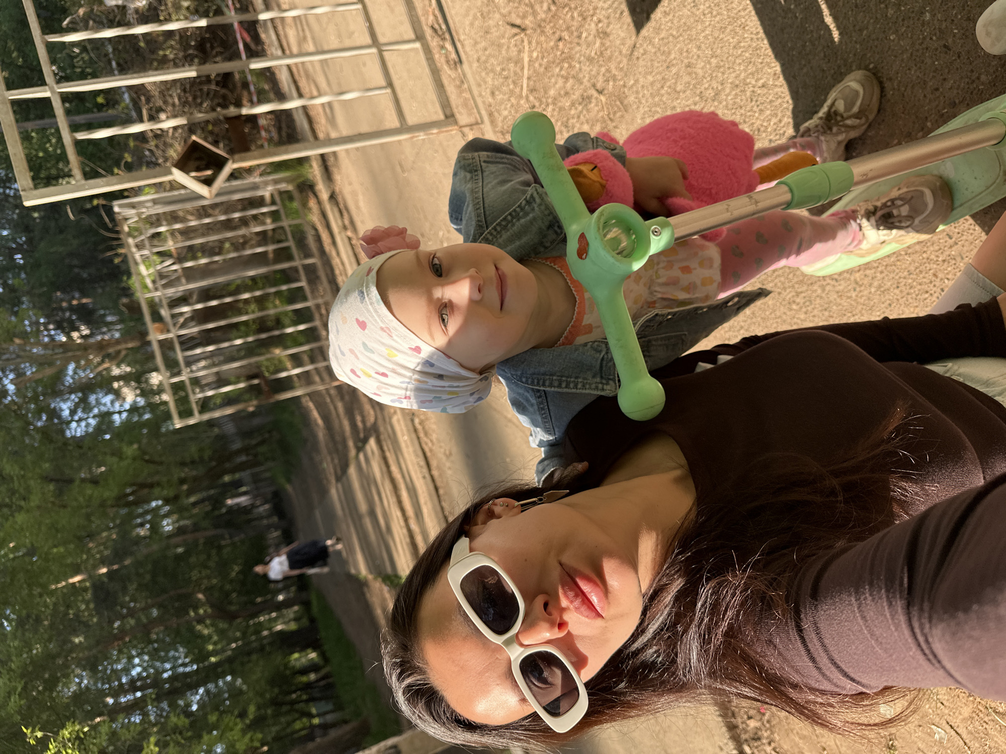 A selfie taken outdoors shows a woman and a child on a scooter. The woman, wearing white sunglasses and a dark top, is in the foreground. The child, wearing a headscarf and a denim jacket, is positioned behind the woman on a light green scooter, holding a pink plush toy. The ground appears to be concrete. In the background, a metal fence and trees are visible. A person is walking in the distance. The location is likely a park or public space in Chaikovsky, Russia.