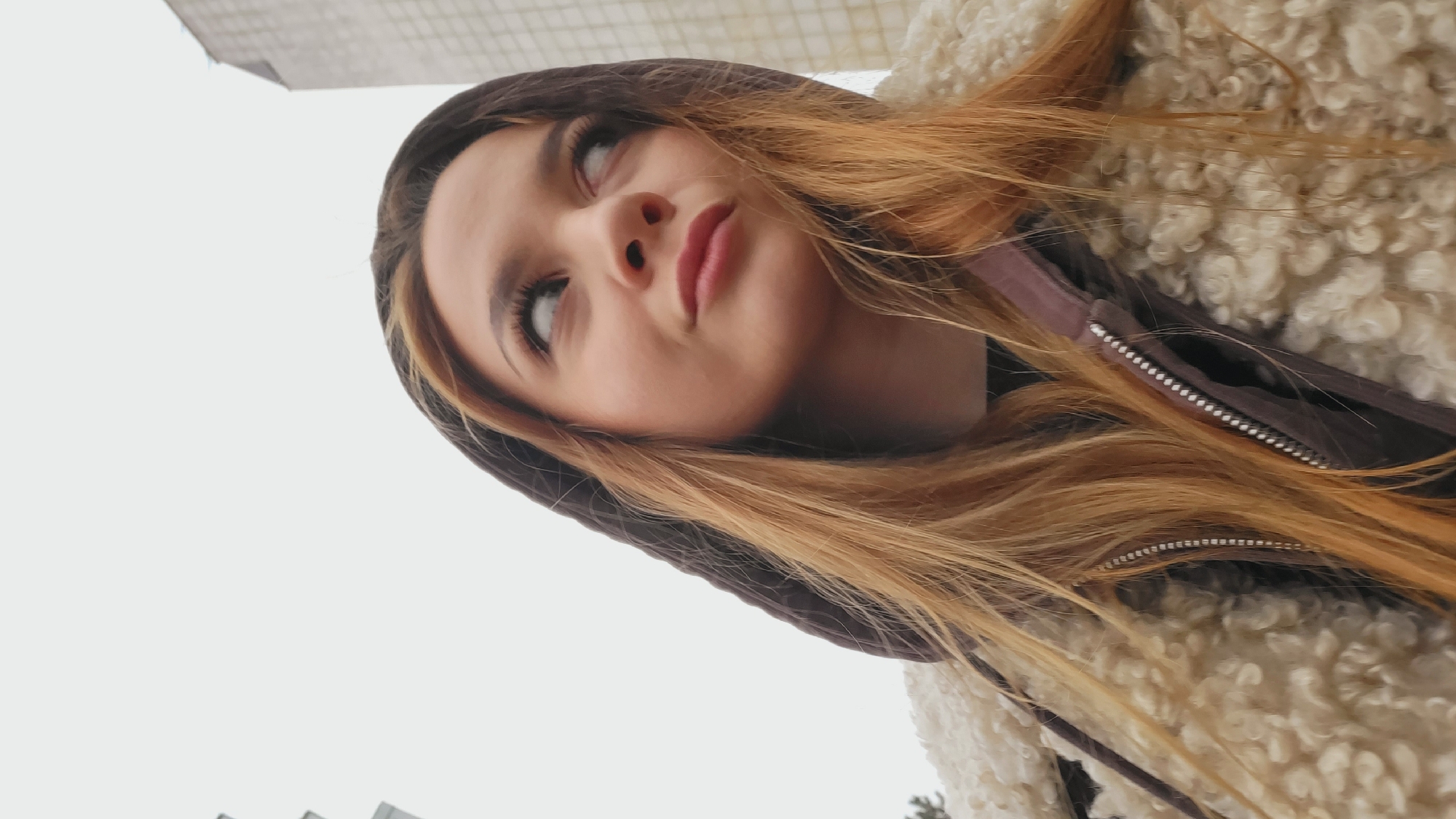 A young woman with light skin, light brown hair with blonde highlights, and wearing a brown hooded jacket with a fluffy white collar, is looking upward and to her left. The photograph is taken from a low angle, with a gray sky in the background and the upper corner of a building visible in the upper left corner. The jacket appears to be zipped up, and the woman's lips are slightly parted. Given the context provided in the instructions, it can be inferred that the location is Chaikovsky, Russia.