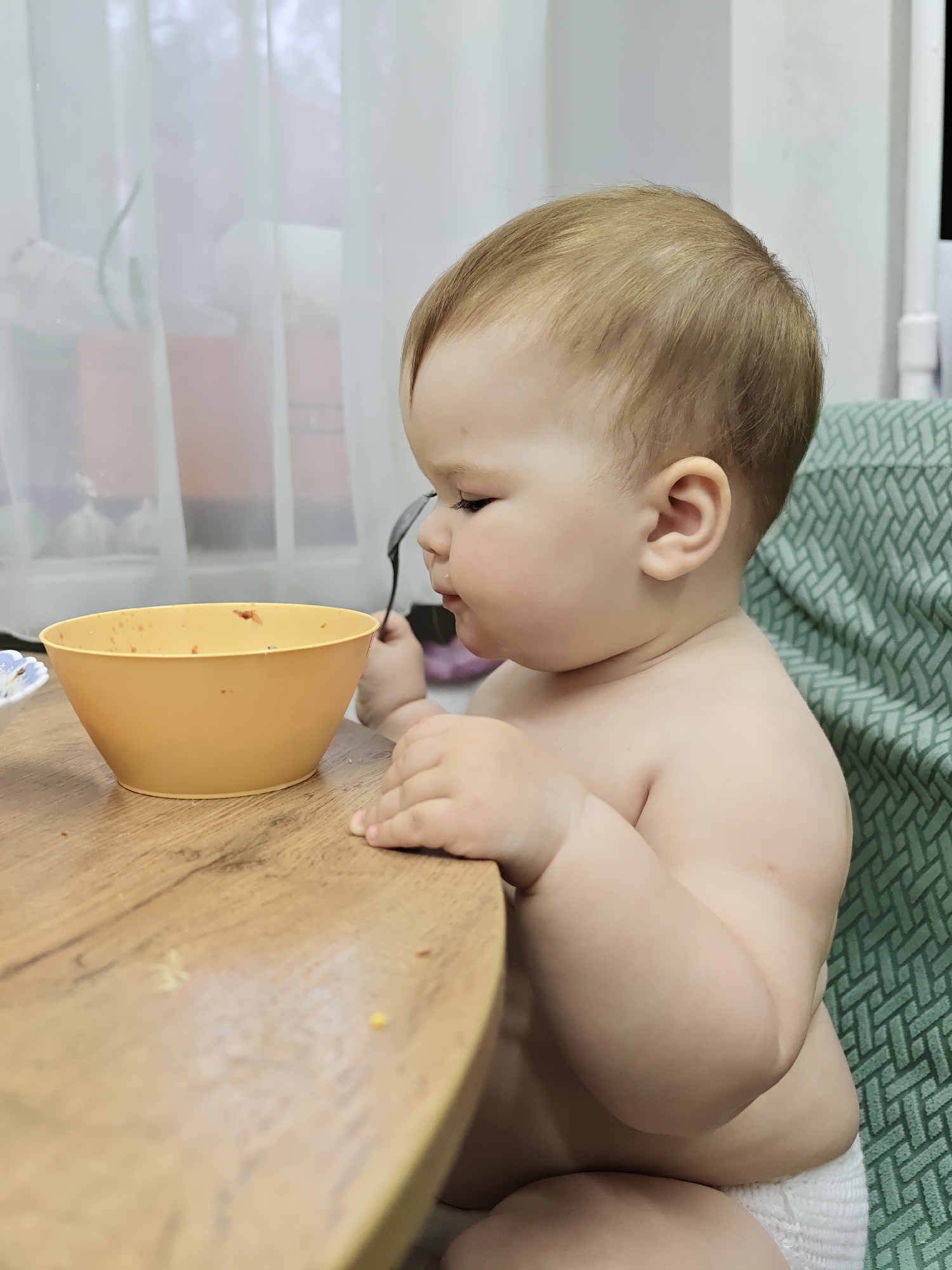 A fair-skinned baby with light brown hair sits at a wooden table next to a yellow bowl holding a spoon to their mouth. The baby sits in a green patterned high chair and wears a white diaper. The background features a window with white curtains. The location is likely a home setting in the city of Chaikovsky, Russia, based on the image's origin. The subject appears to be eating.