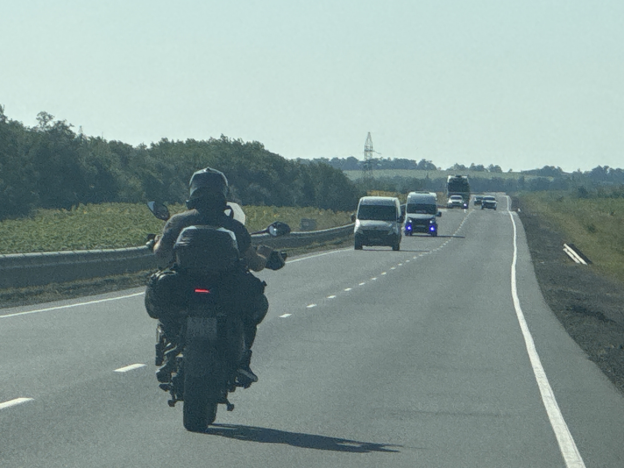 A motorcycle rider wearing a dark helmet and backpack is on a highway in Perevolotsky, Russia. The rider is in the foreground, seen from behind, with the motorcycle's large rear tire and taillight visible. The road has a white dividing line. Several vehicles, including vans and what appear to be trucks, are traveling in the same direction ahead of the motorcycle. The environment is rural, with trees and fields lining the sides of the road. The sky is clear and pale blue. The image appears to be taken from another vehicle on the highway.
