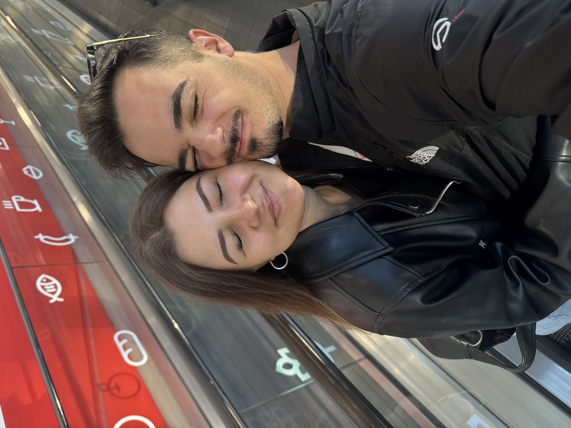 An intimate selfie captures a man and a woman, likely a couple, sharing a tender moment on an escalator or moving walkway. Both individuals have their eyes gently closed and slight smiles, leaning their heads together, conveying affection and contentment.
The man has dark hair and a beard, with sunglasses perched on his head. He wears a black jacket featuring a white "The North Face" logo on the chest and another distinct white logo, possibly "Champion," near the shoulder. The woman has long, dark brown hair, wears a black leather-style jacket, and a silver hoop earring is visible in her left ear.
They are situated on an escalator or moving walkway, suggested by the angled perspective and the visible railings. To their left, a red panel runs alongside, adorned with white line icons typically used to denote food categories in a commercial setting like a supermarket or hypermarket. Visible icons include a banana, a fish, and a loaf of bread. The scene suggests an indoor location, possibly within a shopping center in Sofia, Bulgaria. The overall atmosphere is one of happiness, intimacy, and a relaxed connection between the two people.