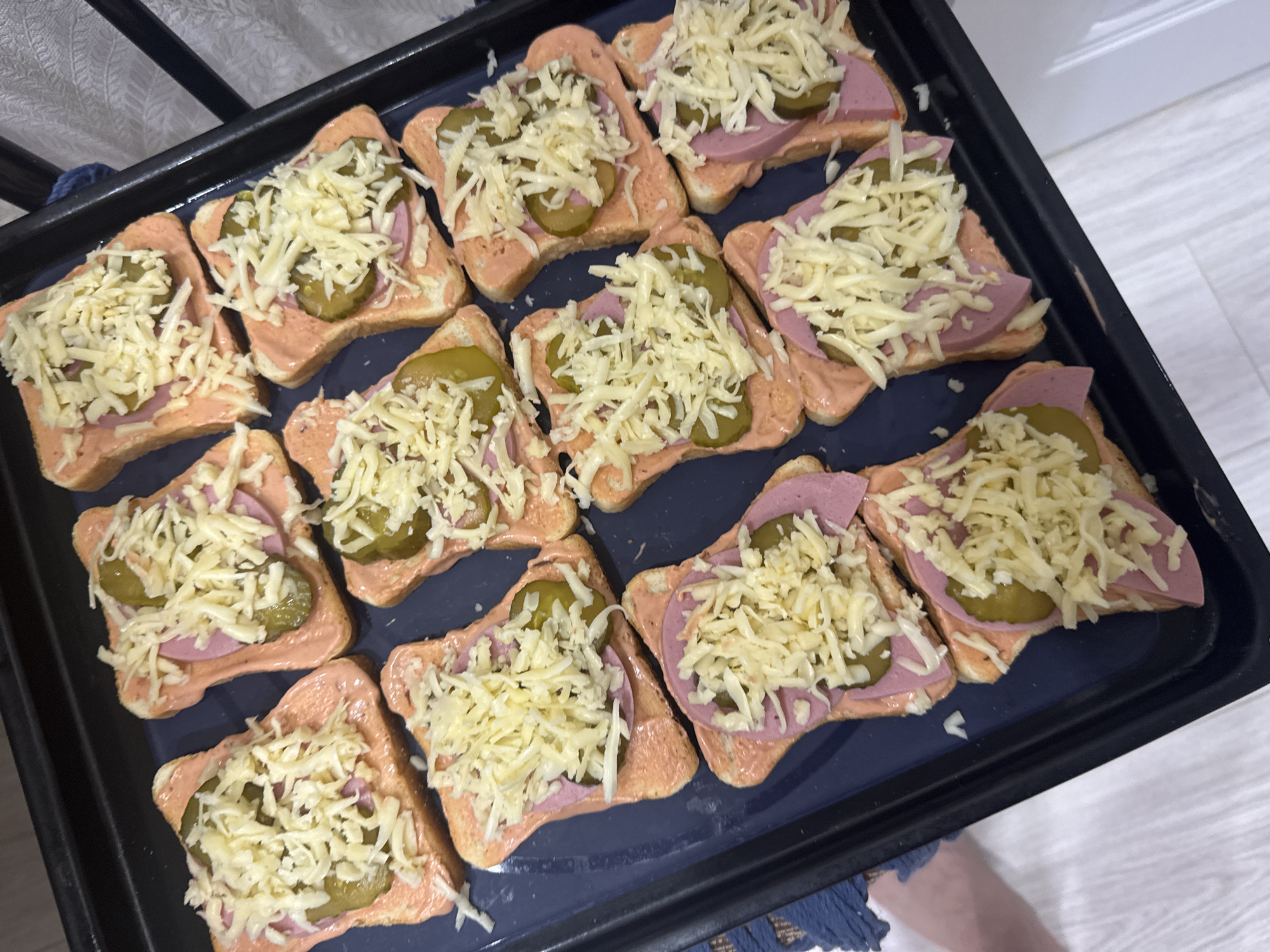 A close-up shot depicts a black baking tray filled with a dozen open-faced sandwiches. Each sandwich consists of a slice of white bread spread with a reddish-pink sauce, topped with a slice of sausage or luncheon meat, sliced pickles, and shredded cheese. The sandwiches are arranged in a rectangular grid on the tray. The tray sits on what appears to be a white surface, possibly a countertop or table. A dark blue fabric is visible in the lower portion of the image. The photo appears to be taken in a residential setting. There is no evidence to suggest that this picture was taken in Izhevsk, Russia.