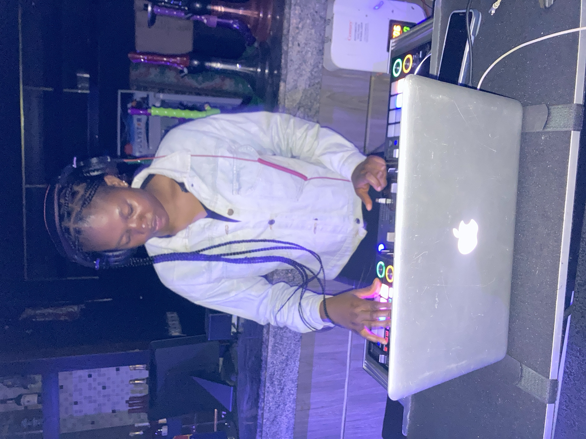 A young woman with braided hair and headphones is standing behind a DJ setup in what appears to be a club in Gwarinpa, Nigeria. She is wearing a white denim jacket and is focused on a laptop and a DJ controller. The room is dimly lit with blue and purple hues, suggesting a nightlife setting. Behind her, there are shelves stocked with bottles and other bar items. Several hookah pipes are visible on a counter to her right. The overall atmosphere is that of a lively entertainment venue.