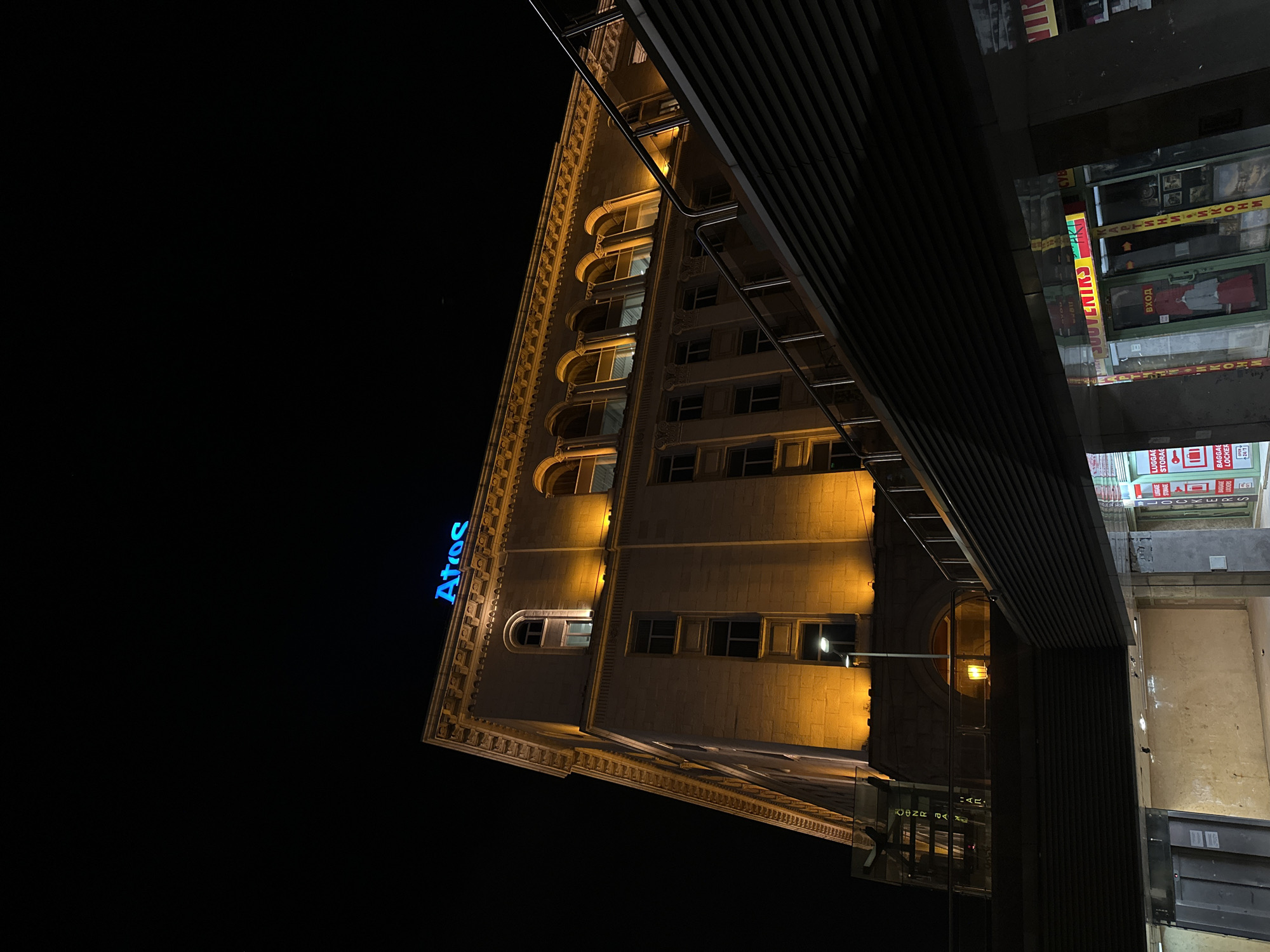 The image captures a nighttime view in Sofia, Bulgaria, showcasing the Central Department Store (TZUM). The building, illuminated with yellow lighting, has an "Atos" sign in blue neon lights. A modern, dark-colored structure, possibly a pedestrian bridge or part of a neighboring building, overlays the scene. Stores and signs are visible in the lower-right, indicating commercial activity. The sky is dark and devoid of detail, indicating night.