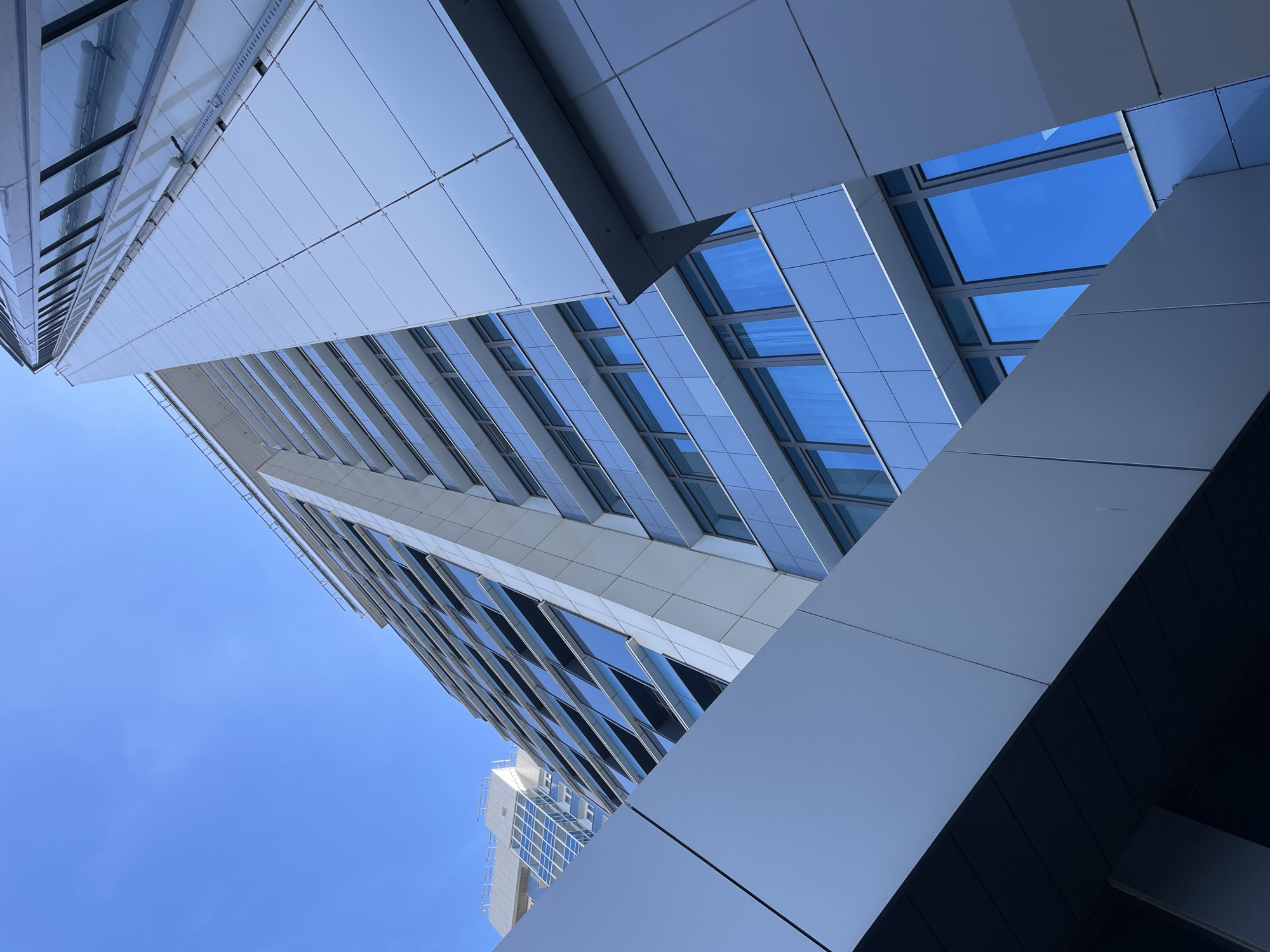 The image shows a low-angle view of modern architecture, presumably a building exterior. The facade consists of a combination of glass windows, steel, and aluminum panels arranged in a grid-like pattern. The buildings are illuminated by sunlight. The sky is clear and blue. The buildings have various shapes and edges. I don't have enough information to determine the location within Krasnoyarsk, Russia.