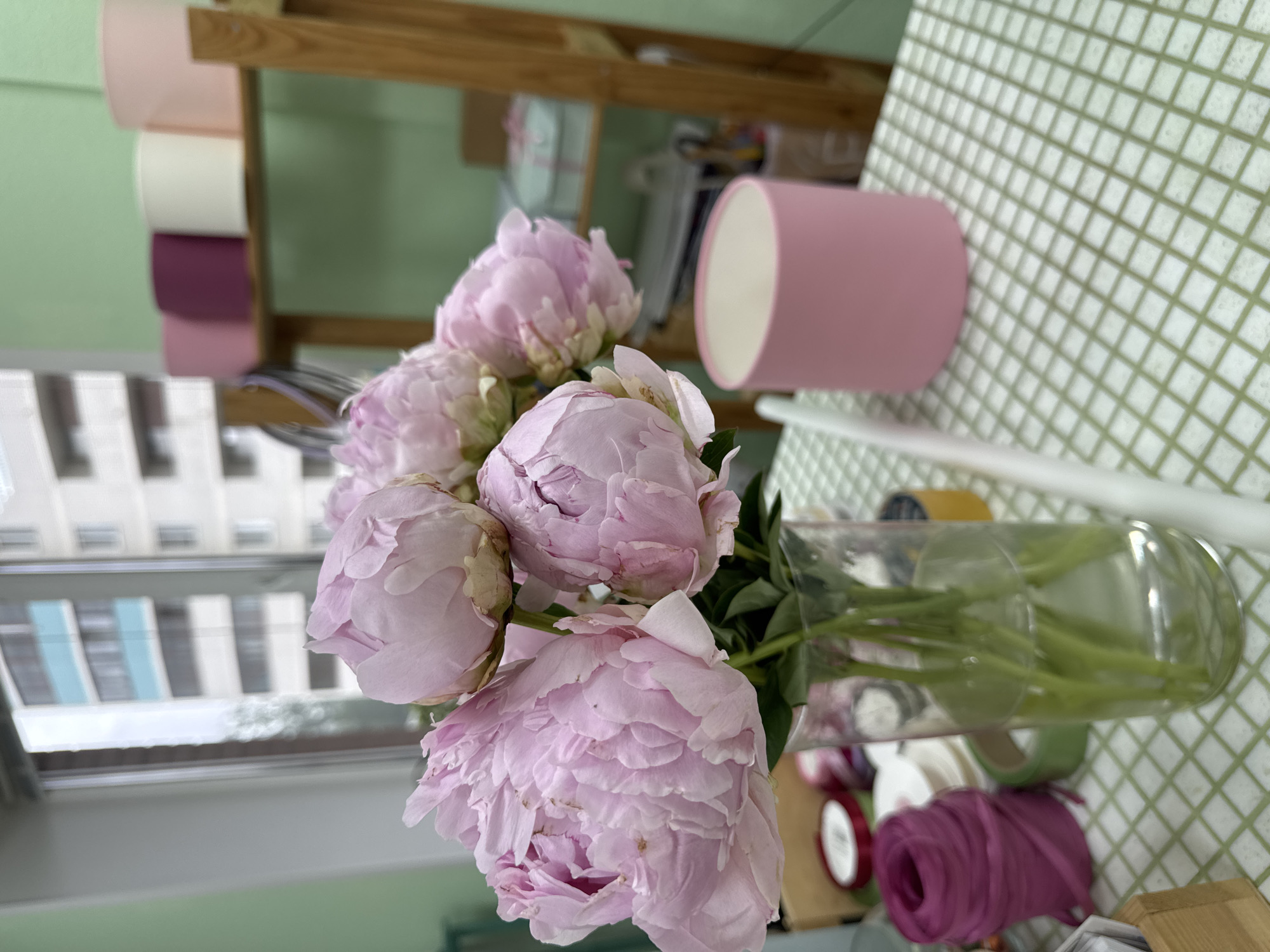 The image shows a bouquet of light pink peonies in a clear glass vase. The vase is sitting on a surface covered with a green and white checkered pattern. A pink cylindrical object is next to the vase. In the background, a wooden shelf holds rolls of material in various colors, including pink, white, and purple. A window is visible to the left, showing a building with alternating light and dark-colored sections. Based on the available information and the style of the building in the window, I am unable to confirm the location as Izhevsk, Russia. Additional details are needed to accurately determine the location.