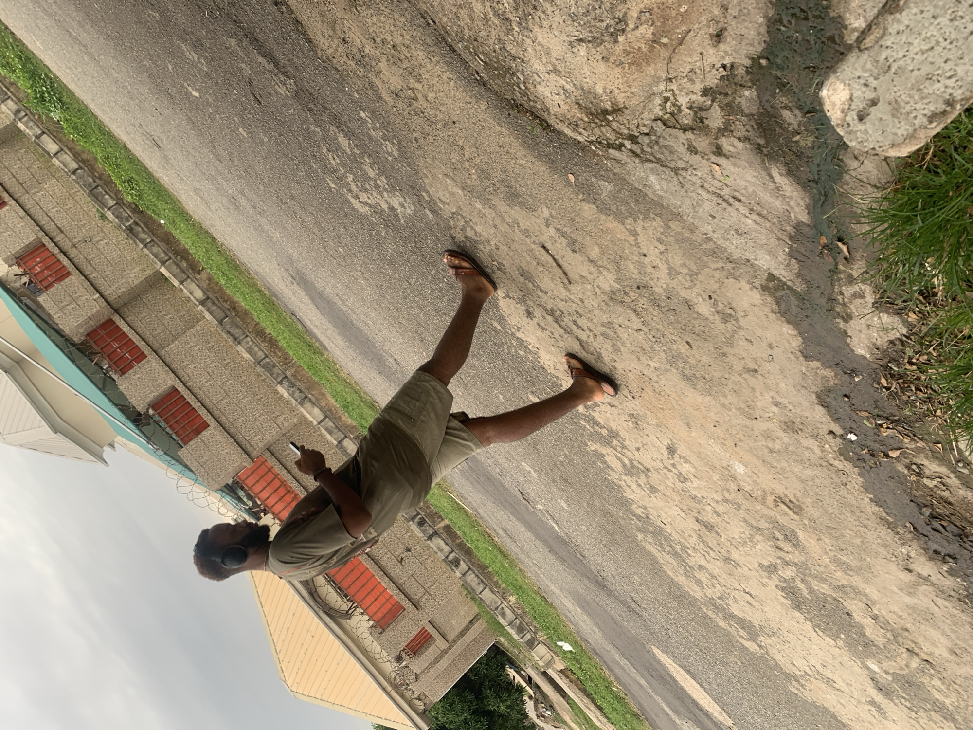 A young African man with dark skin, a beard, and short dark hair is standing on a paved road. He is wearing a green t-shirt and khaki shorts. He has black headphones on his ears and is holding a smartphone in his right hand. He is barefoot, wearing brown flip-flops. The man is looking down at his phone. Behind him, there is a building with a light-colored roof and a textured stone facade. The building has red-framed windows and barbed wire fencing. The area appears to be an urban environment, possibly a street in Abuja, Nigeria, given the architecture and vegetation. The road is asphalt and shows signs of wear and tear, with patches of dirt and water stains. Green grass grows along the side of the road. The sky is overcast.