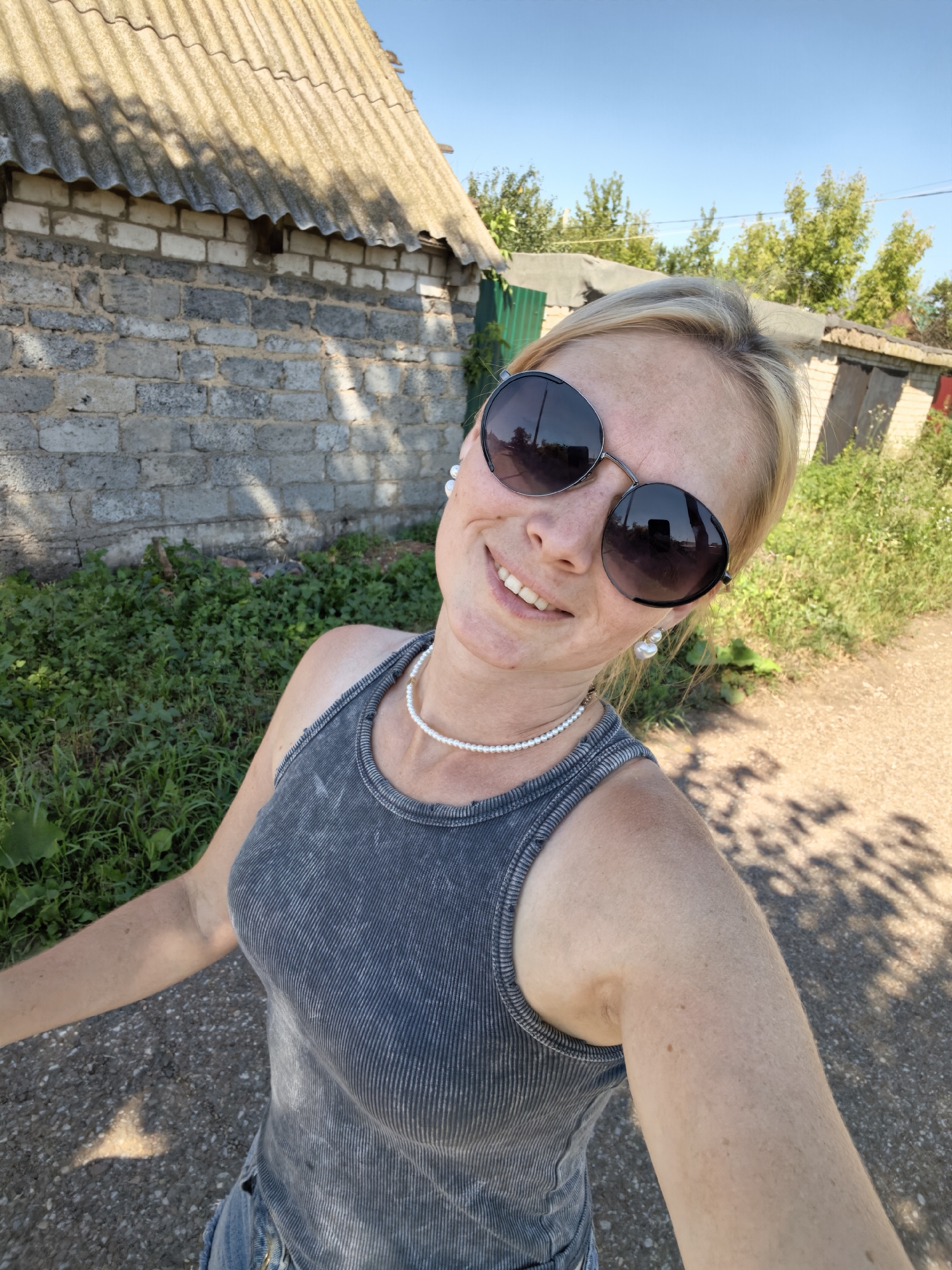 A blonde woman wearing large, dark sunglasses, a grey ribbed tank top, and a pearl necklace poses for a selfie. She is standing outdoors on a dirt road, with a brick building and overgrown greenery in the background. The sun is shining, casting shadows on the ground. The image appears to have been taken in a rural or suburban area. The location in the city of Sterlitamak, Russia cannot be determined from the visual information provided.