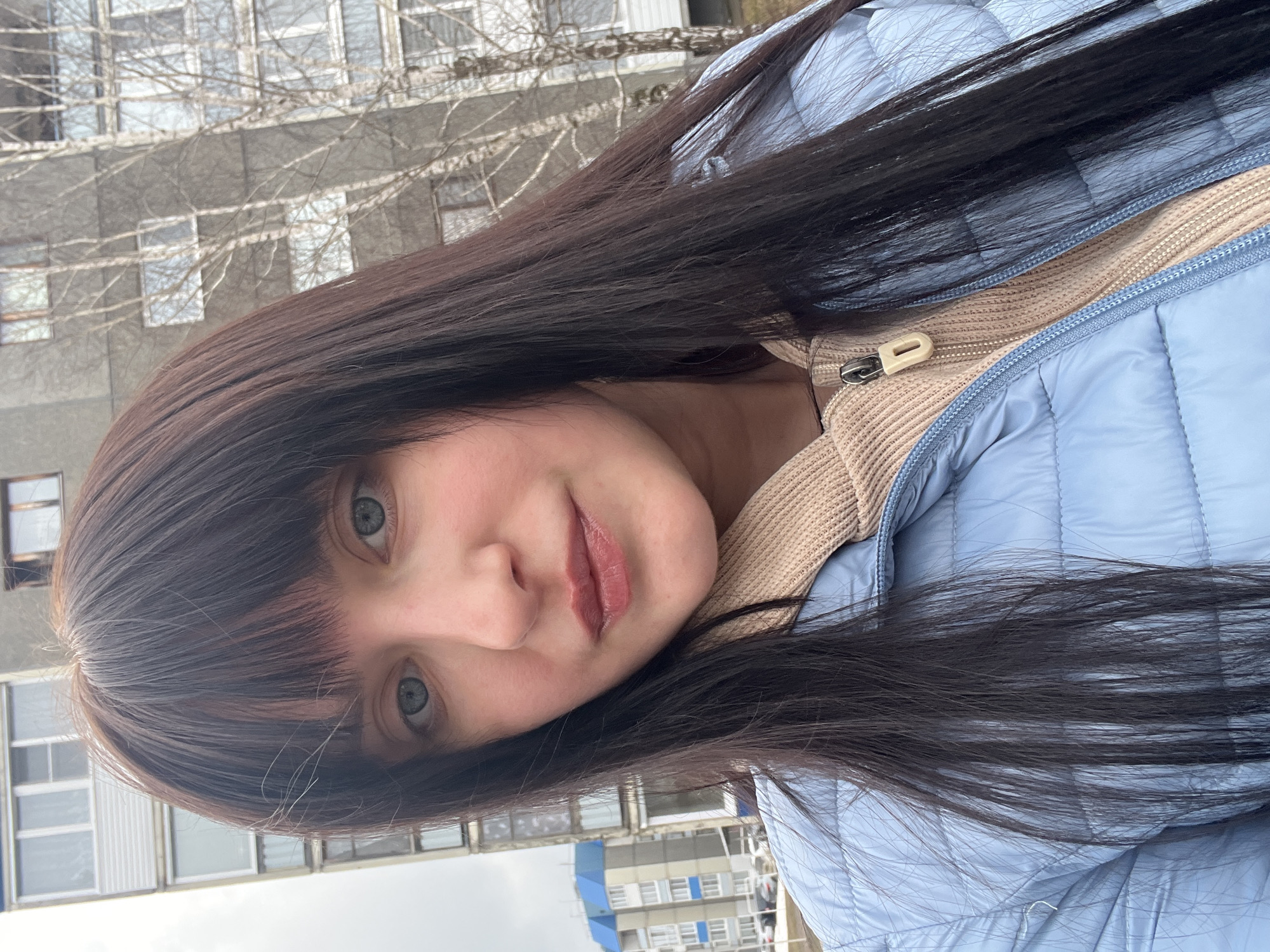 A young woman with long, dark hair and bangs is pictured in a selfie. She wears a light blue quilted jacket over a beige sweater or top. She has fair skin and visible makeup on her lips. Behind her is a multi-story building with numerous windows. The location is likely in the city of Марковский, Russia. The sky is overcast. There is a bare tree branch visible near the building.