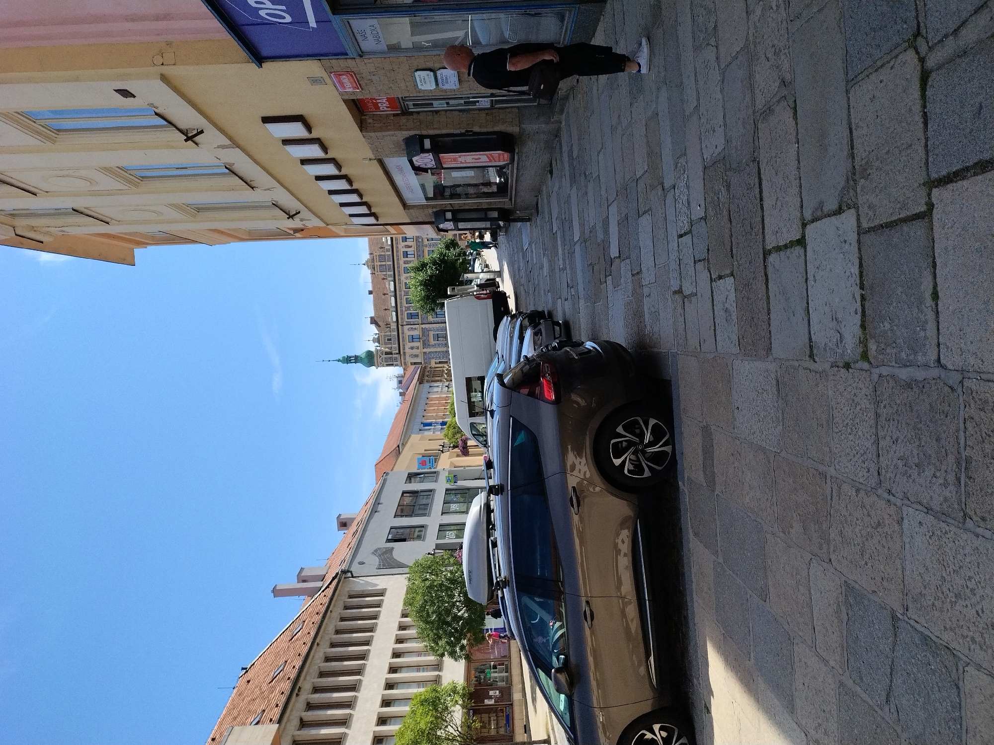 The image depicts a street scene in Písek, Czechia, on a sunny day. The perspective is from a pedestrian's eye level, looking down a cobblestone street lined with buildings. In the foreground, a brown station wagon with a roof box is parked. Further down the street, several other cars are visible.
On the right side of the street, a man wearing a black polo shirt and black pants with white sneakers stands facing a shop window. The shop has signage in Czech, including "NASE NABIDKA" (Our Offer). The building itself is a light yellow color.
On the left side of the street, there are buildings with tiled roofs and multiple windows. Green trees line the sidewalk in front of these buildings. In the distance, an architectural landmark with a green spire is visible against a clear blue sky. The street pavement is made of large, irregular stone blocks.