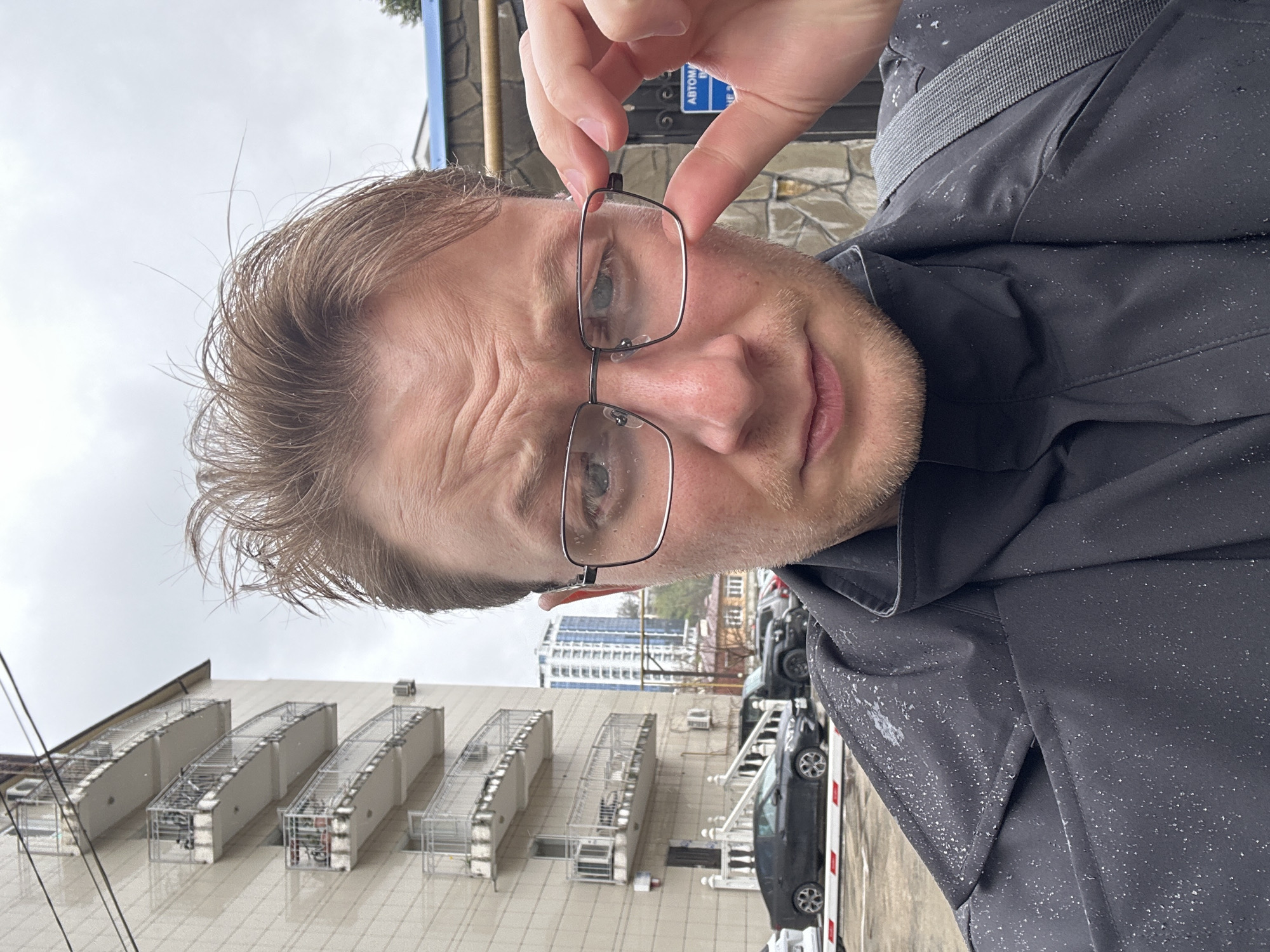 A medium close-up, slightly tilted photo shows a man holding a pair of rectangular metal-framed glasses in front of his face. He has light-colored hair, fair skin, and blue eyes. The man is wearing a dark gray or black jacket.
Behind the man, the background is partially in focus and suggests an urban environment. There is a large, light-colored building with several protruding structures that could be air conditioning units. Cars are visible in the background.
The location is Sochi, Russia, as indicated by a partially visible sign above the man's hand. The sky is overcast and the overall scene suggests a cloudy or rainy day, as there are visible water spots on the man's jacket and the pavement.
