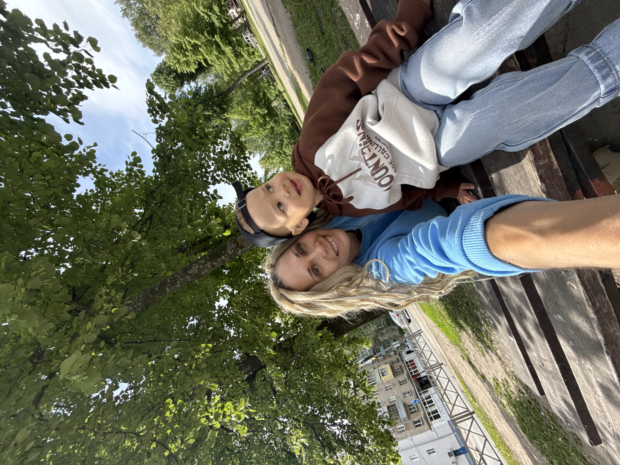 A selfie shows a blonde woman with long dreadlocks, wearing a blue sweater, lying on a park bench with a young child. The child is wearing a brown and white hoodie and a baseball cap. Both are looking at the camera and smiling. The bench appears to be wooden, and is located in a park. Trees with green leaves are visible in the background. Beyond the trees are buildings, indicating that the park is located in an urban environment. Based on the style of the buildings and general appearance of the park, it is possible this photograph was taken in Rusanovo, Russia.