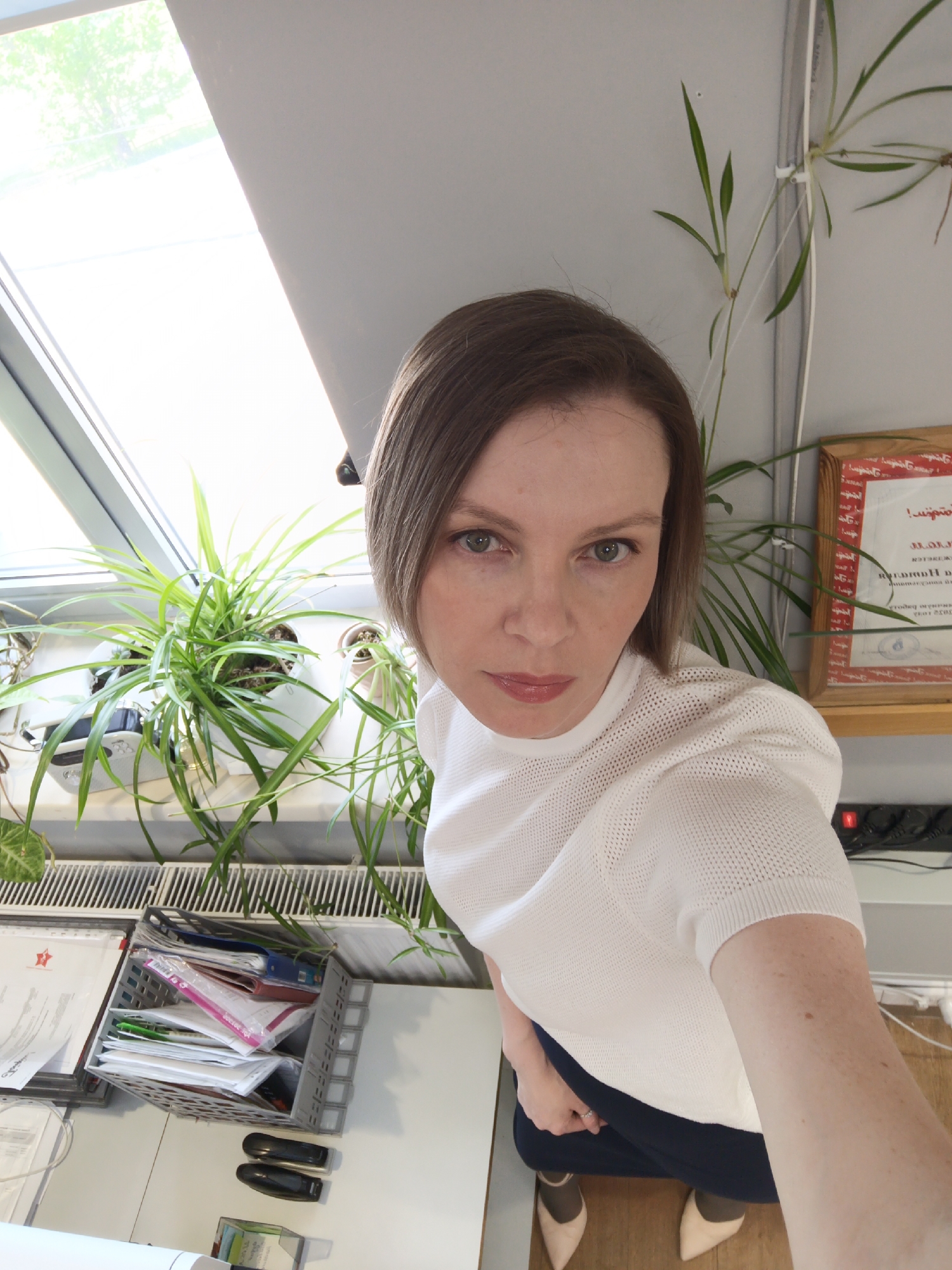 A woman with short brown hair, wearing a white shirt and dark pants, is taking a selfie. She is indoors, next to a window with plants on the windowsill. A file basket and staplers sit on a table next to her. A certificate hangs on the wall behind her, next to a plant in a vase. She is wearing beige high heels. The certificate likely indicates the location is Chaikovsky, Russia.