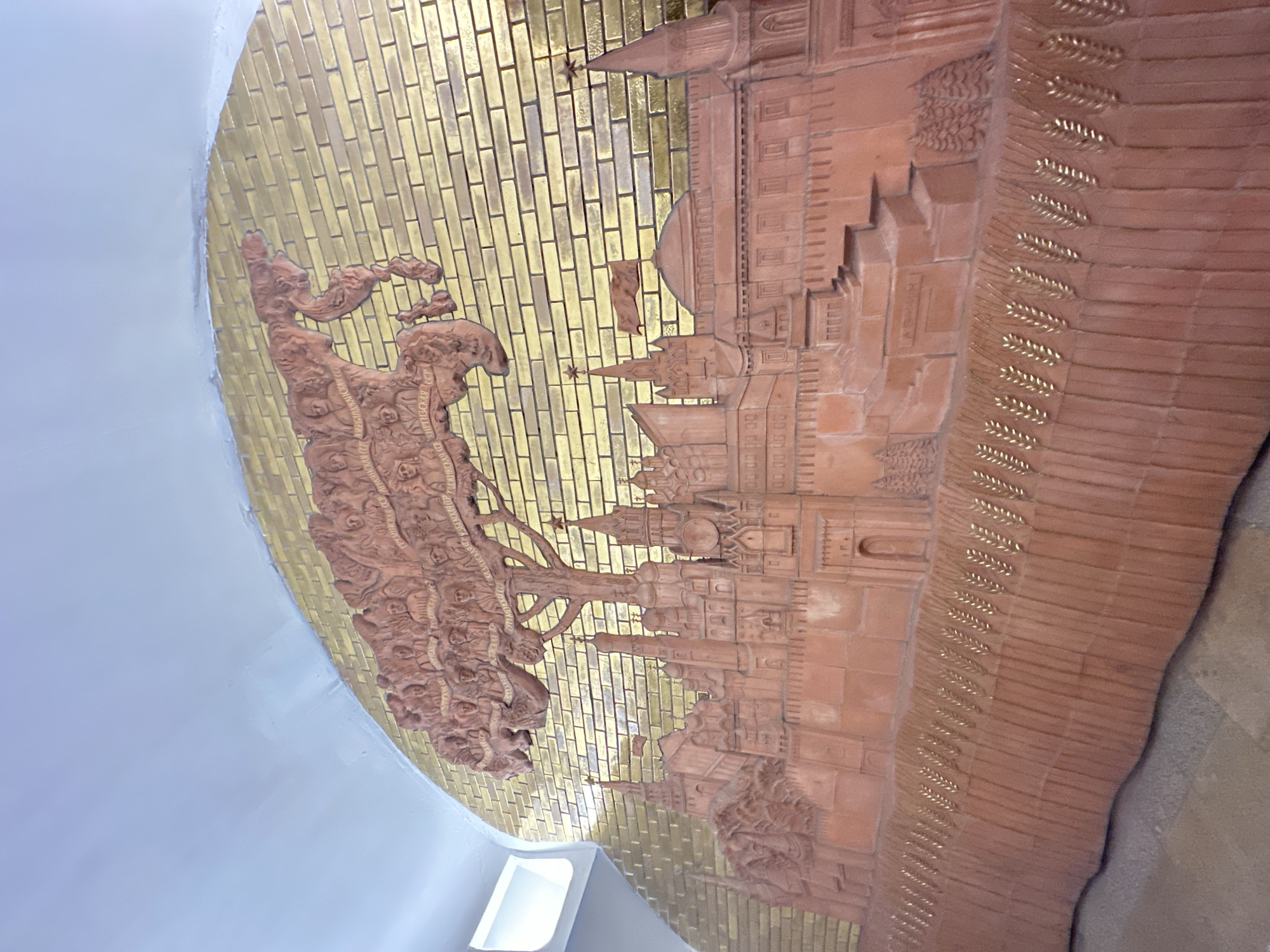 The image captures a portion of a decorative panel or mosaic within the Moscow Metro in Russia. The panel is curved, suggesting it is part of a tunnel wall or ceiling. It features a relief-like depiction of the Moscow Kremlin and other historical landmarks, executed in a reddish-brown material. A tree-like structure is depicted on the left, featuring several figures, possibly representing people or historical events. The background is made of golden-colored, brick-like tiles. The surrounding structure is painted in a light-colored paint.