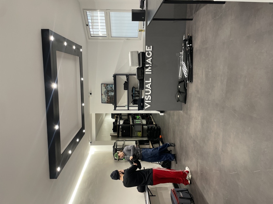 The image depicts an indoor commercial space, likely an equipment rental facility or studio for visual media, observed from a slightly elevated perspective. The room is well-lit with artificial lighting.
In the foreground, two men are standing on a dark grey, possibly concrete or tiled floor. The man closest to the viewer is seen from behind, wearing a black beanie, a black long-sleeved top, and red athletic pants with white stripes down the sides. He appears to be holding a small black object, possibly a camera or phone, and is looking towards the second man. The second man, positioned slightly further into the room and facing the first man, has short hair and is wearing a grey long-sleeved shirt, dark blue pants, and a green baseball cap. He is looking down at something he is holding in his hands, which also appears to be black equipment. A small black bag is slung over his shoulder. Their interaction suggests they are discussing or inspecting equipment.
To the left of the men, a large, dark-colored rectangular frame is mounted on the white wall. This frame has numerous bright, circular lights embedded along its inner edges, providing additional illumination. Below this frame, a thin, linear strip of light is visible along the wall.
Further into the room, a prominent dark grey counter or desk runs horizontally across the space, separating the area where the men are from the background. Vertically displayed in white uppercase letters on the front of this counter are the words "VISUAL IMAGE". On top of the counter, several black bags and possibly some grey cushions are stacked. To the right of the counter, several black light stands and other equipment are neatly arranged or stacked.
Behind the counter, a series of black metal shelving units are visible, filled with various black cases, bags, and other equipment, indicating a storage or display area for rental gear. High up on the back wall, above the shelving and counter, is a rectangular window with horizontal blinds, allowing some natural light into the space. A security camera is mounted on the white wall above the shelving units, and a framed picture or poster hangs on the wall below it, to the left of the shelves. The overall impression is one of a functional and organized professional environment for visual production equipment.