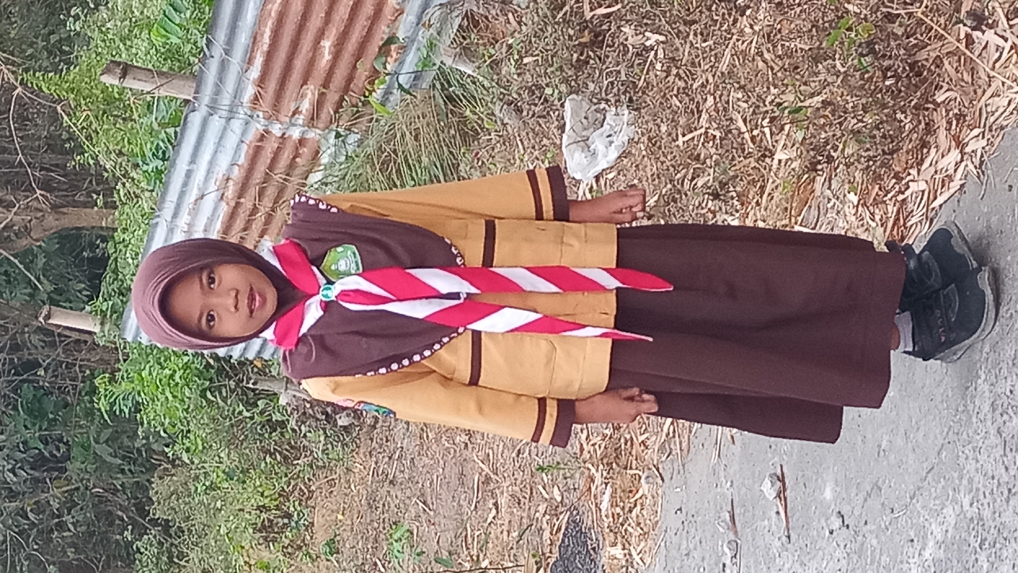 A young Indonesian girl, appearing to be a scout, stands outdoors. She is wearing a brown hijab, a tan uniform jacket with brown trim, and a brown skirt. A red and white striped scarf is draped around her neck, fastened with a circular badge. She wears dark sneakers. The background consists of dry vegetation, some trees, and a corrugated metal barrier on the right. The ground is a mix of dirt and a paved path. The lighting suggests it is daytime. The image is taken in Pare, Indonesia.