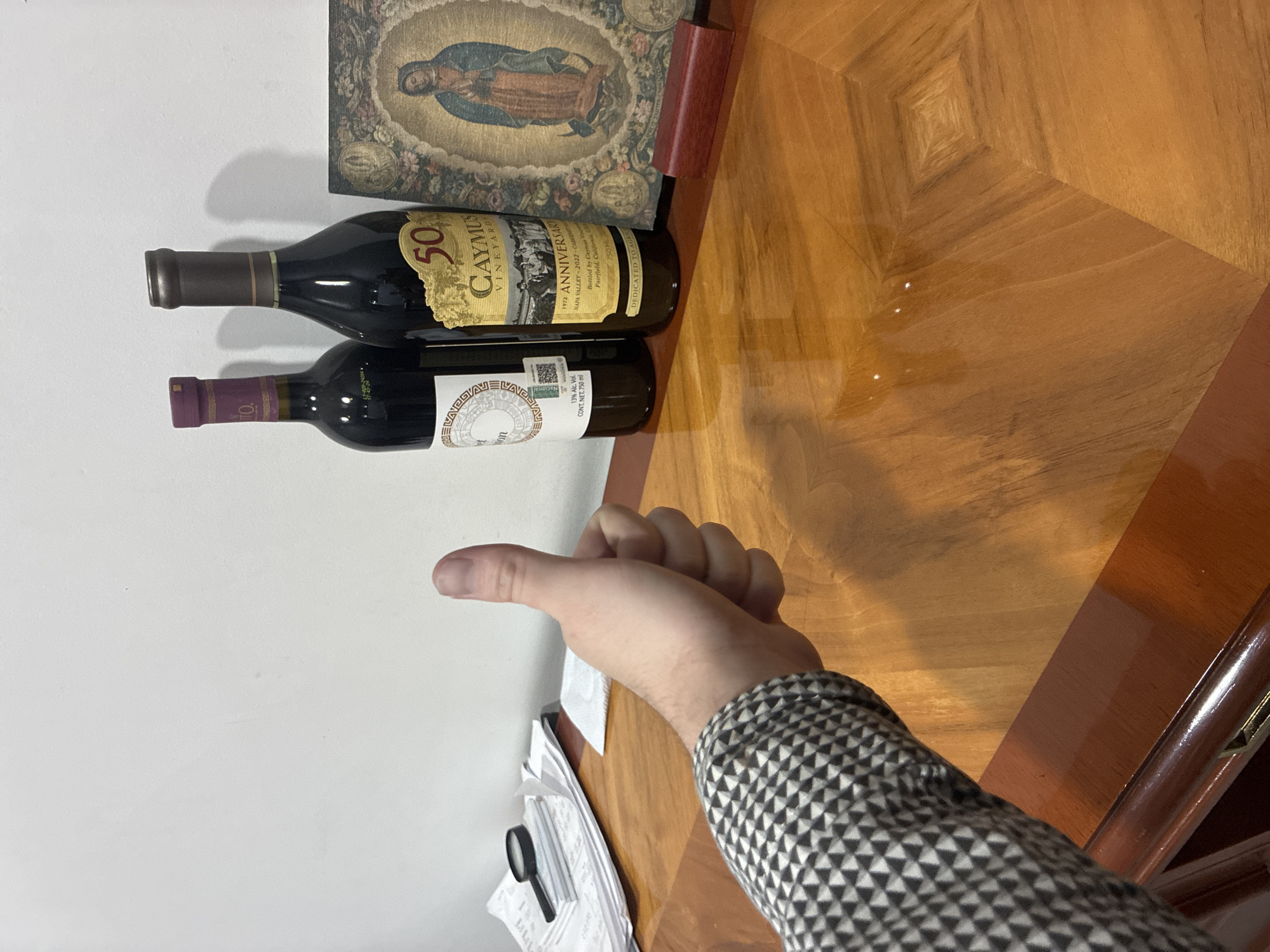 The image depicts two bottles of wine lying on a wooden surface next to a framed religious painting. The top bottle is labeled "Caymus Vineyards 50th Anniversary Napa Valley." A person's hand with a thumb extended upward is partially visible. The location is Cuautitlán Izcalli, Mexico. Other objects present include papers, a magnifying glass, and a decorative sleeve on the arm of the person.