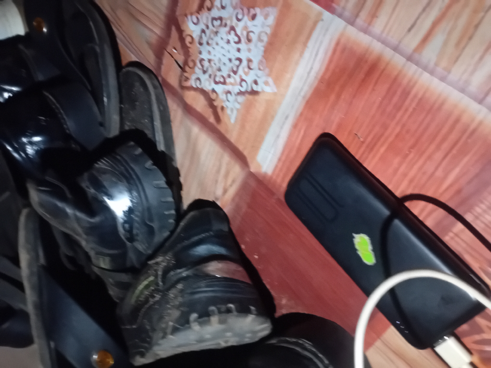 The image shows a collection of black footwear, including boots and sandals, piled on a wooden floor. A black power bank with a green leaf-like sticker is charging with a white cable. To the right, a piece of paper with a star-shaped stencil design is visible on the floor. The scene is dimly lit. The location is Unknown, Unknown.